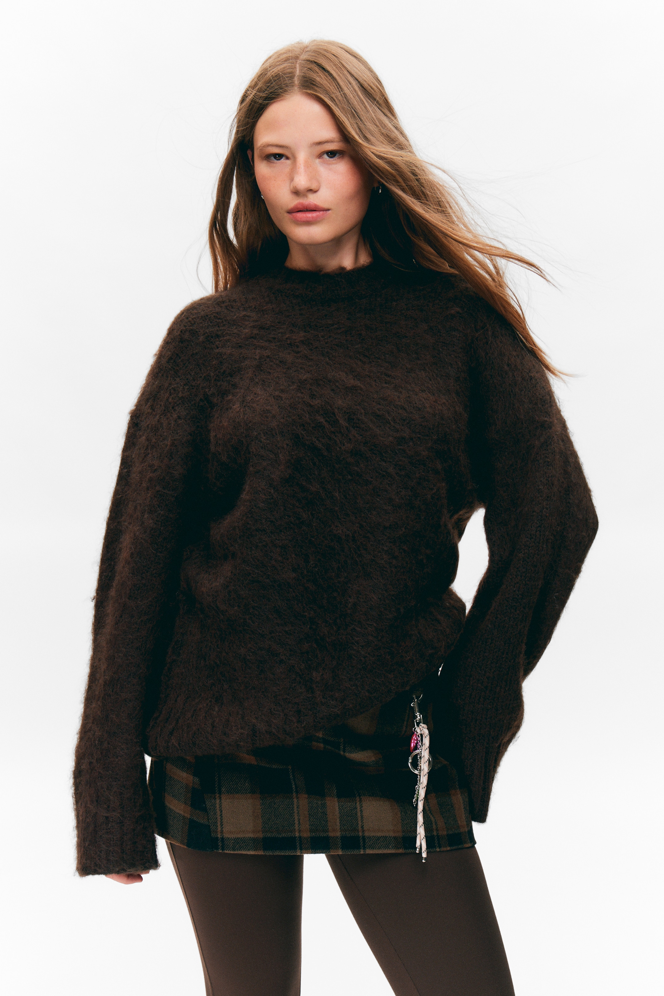 Visa större bild: En mörkbrun luddig oversized tröja presenteras från en framåtvänd vinkel, med en rund hals och långa ärmar. Den är lagrad över en brun och grön rutig skjorta, med en silverfärgad nyckelring som hänger från dess högra sida.