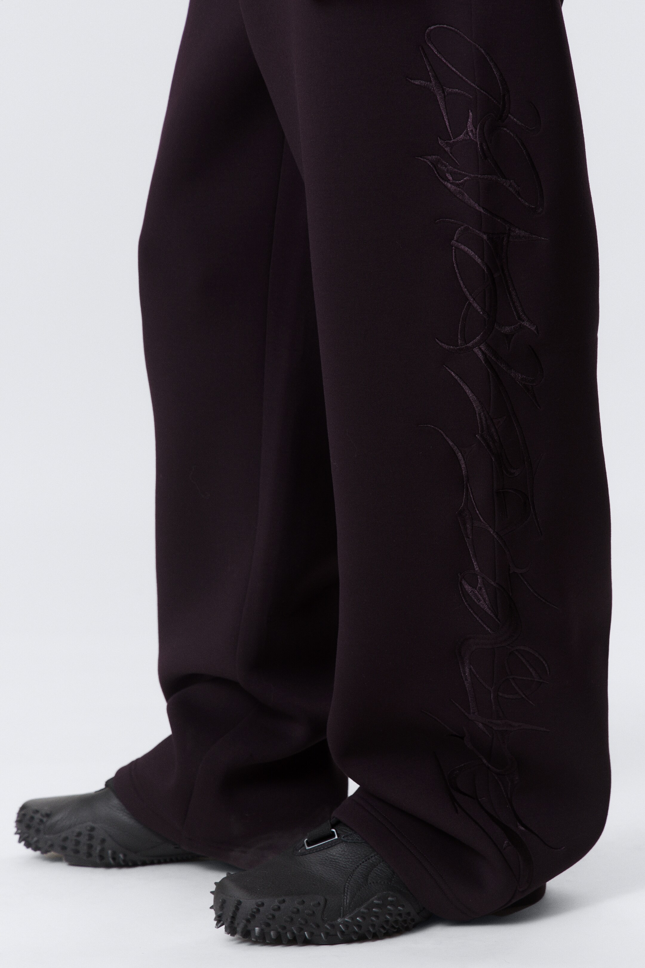 Agrandir l'image: Des pantalons ample de couleur violet foncé présentent un design brodé complexe, ton sur ton, qui court verticalement le long du côté extérieur de la jambe, s'étendant vers l'ourlet.