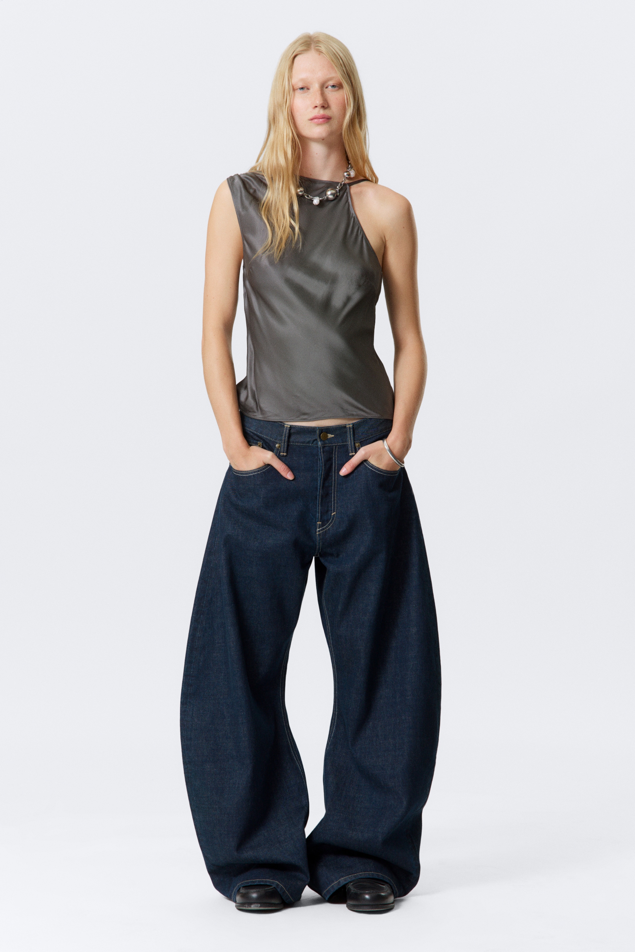 View larger image: A dark gray sleeveless top with an asymmetric neckline, featuring a wide left strap and a right shoulder cut-out, is being worn by the model and styled with dark wash wide-leg jeans.