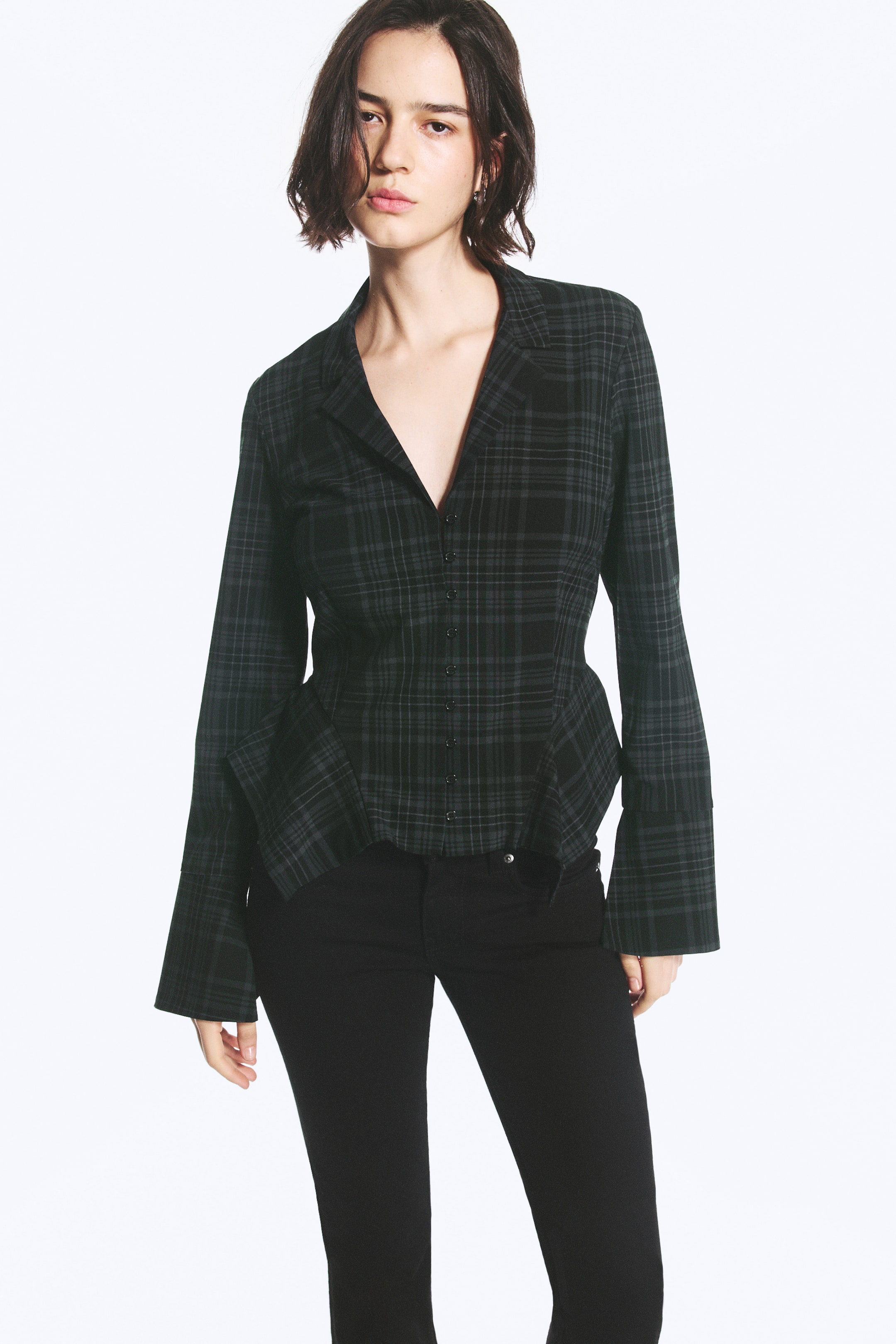 Visualizza immagine più grande: Una camicia a linea A in un motivo tartan scuro è indossata dalla modella, con chiusura frontale con bottoni, rever a lancia e maniche lunghe con ampi polsini allungati. La camicia presenta un orlo svasato e strutturato ed è abbinata a pantaloni neri.