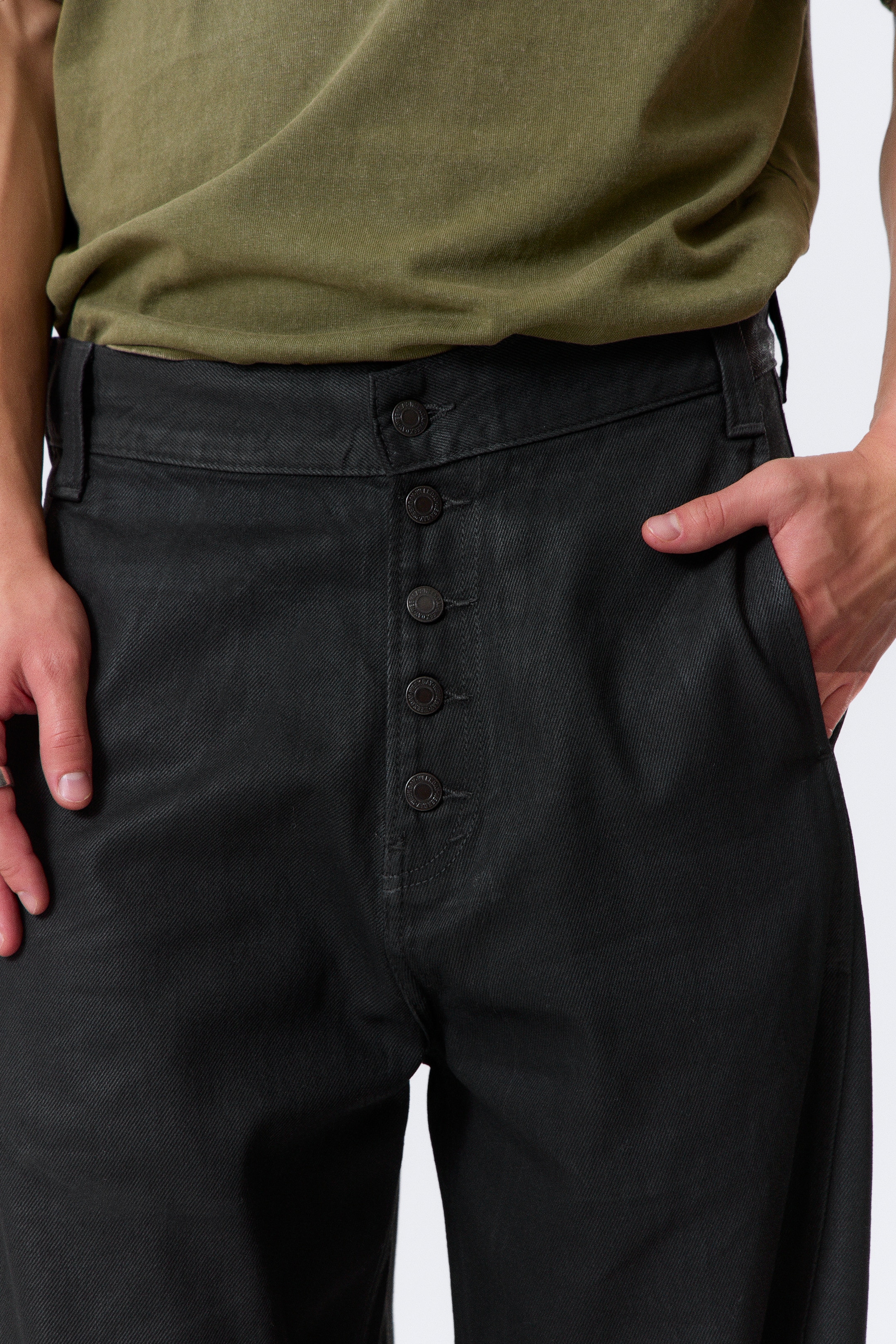 View larger image: Black twill pants are being presented from a frontal view, featuring a five-button fly with exposed dark buttons down the center front. The pants are a relaxed fit with side pockets, and an olive green t-shirt is being tucked into the waistband.