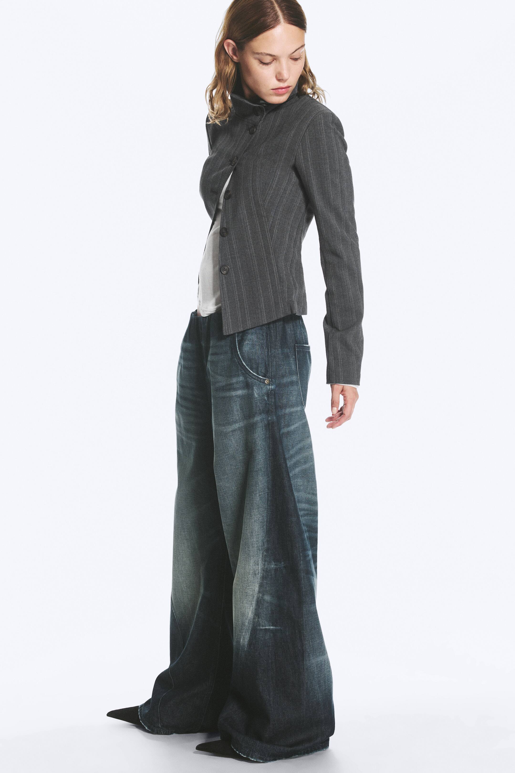 Agrandir l'image: Un jean style jogger à taille basse et coupe ample, délavé bleu foncé, porté par le mannequin, avec une taille élastiquée, des découpes incurvées et des poches latérales en biais avec détails rivetés. Le jean est associé à une veste grise à fines rayures et à des chaussures noires à bout pointu.