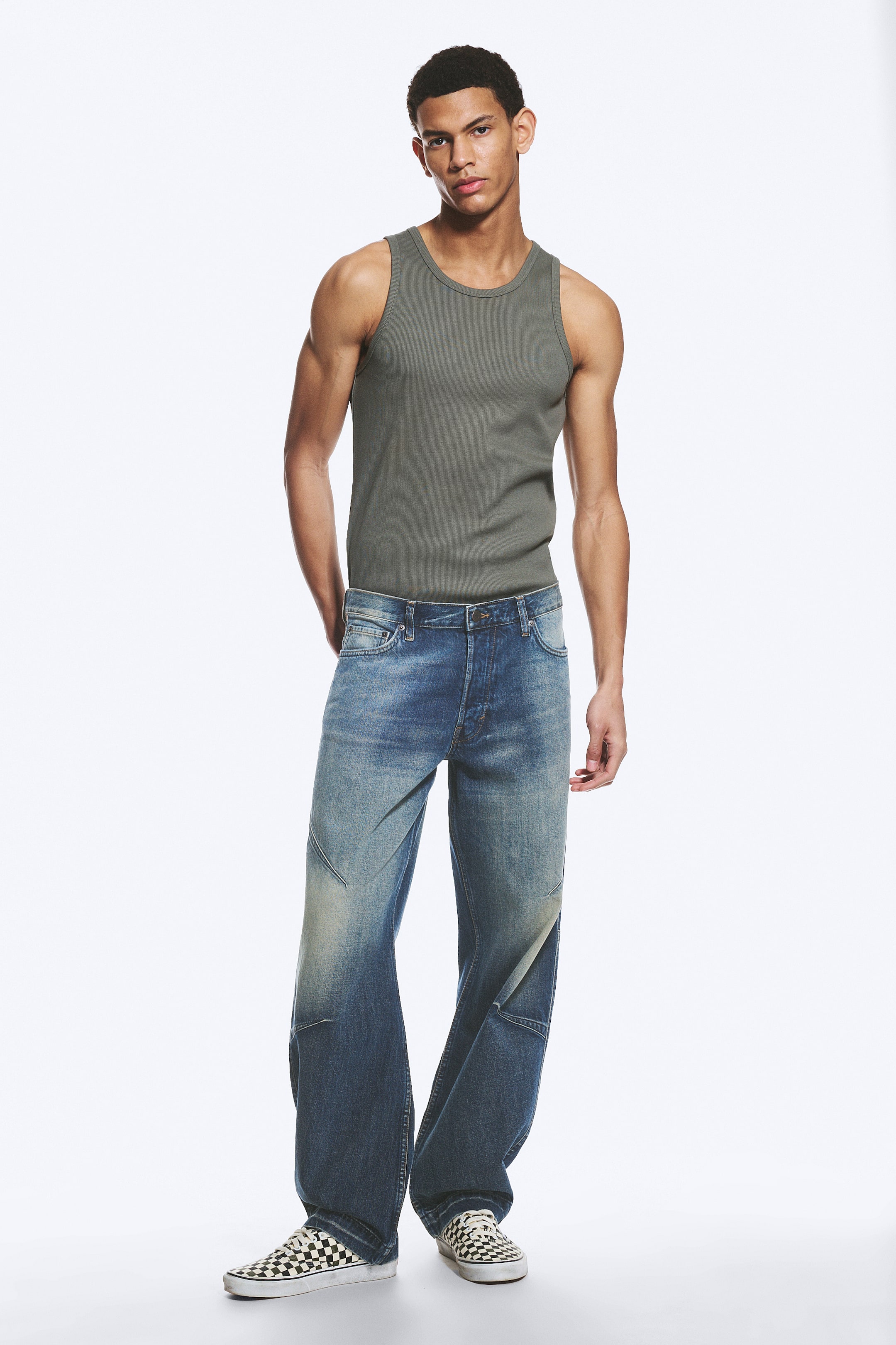 Se større billede: En olivengrøn ribbet tanktop med rund hals og ærmeløst design, stukket ned i blå jeans med brede ben. Jeansene viser forskellige vaskenuancer og slidte detaljer, hvor de brede ben hviler over ternede slip-on sko.