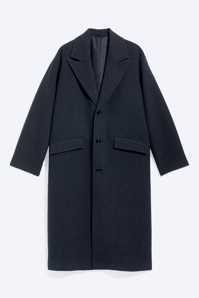 à apresentado um casaco comprido, azul-escuro, de abotoamento simples, visto de frente, com uma silhueta oversized, lapelas de entalhe marcadas e fecho frontal de três botões. Foi concebido com dois bolsos embutidos com aba e bainhas com acabamento limpo, destacando o seu tecido texturizado em mistura de lã. - 1