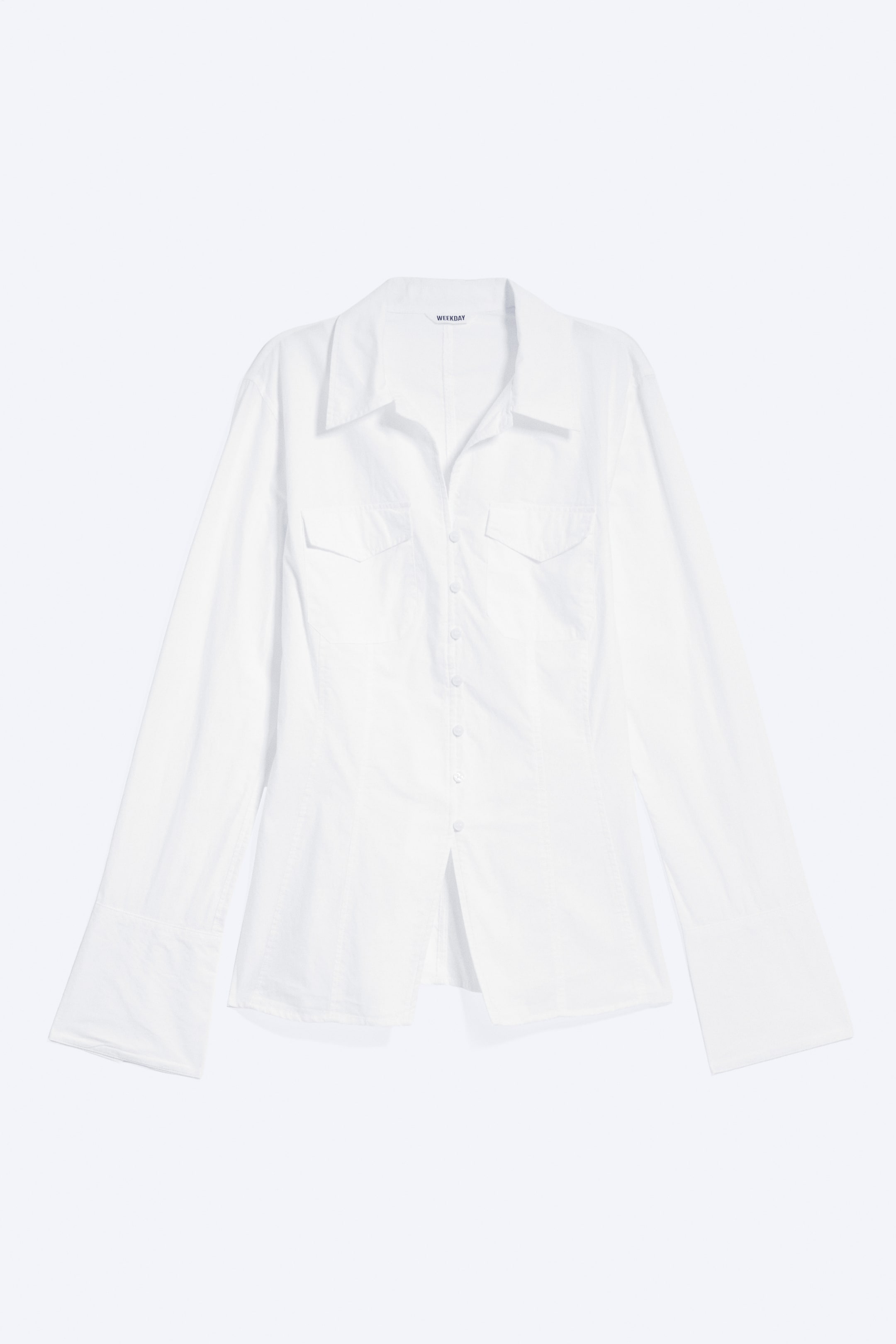 Agrandir l'image: Une chemise blanche est présentée de face, avec un col pointu, une patte de boutonnage intégrale avec des boutons clairs et deux poches plaquées à rabat sur la poitrine. Elle présente des manches longues avec de larges poignets et un ourlet légèrement évasé.