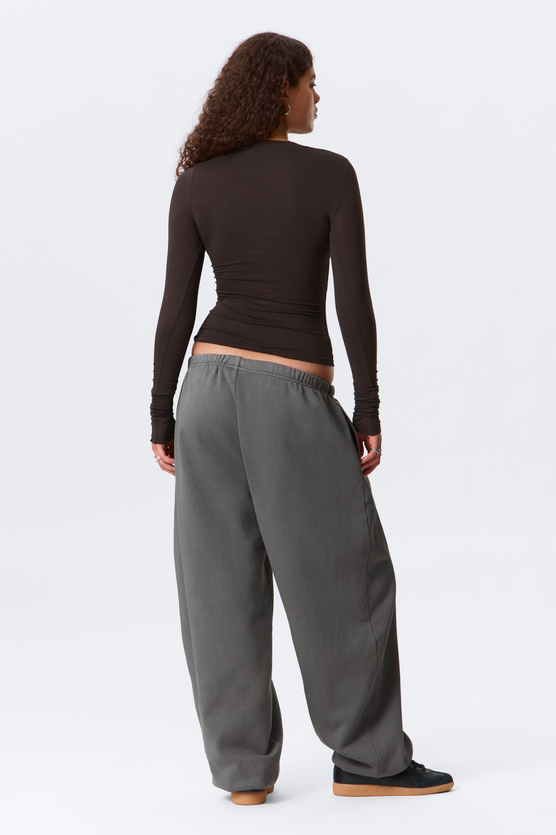 Agrandir l'image: Un haut à manches longues brun foncé est présenté de dos, montrant sa silhouette ajustée et ses longues manches avec des poignets froncés. Un pantalon de survêtement ample gris foncé avec une taille élastique est porté bas sur les hanches, avec des jambes larges qui se rassemblent aux chevilles sur des baskets noires avec des semelles brunes.