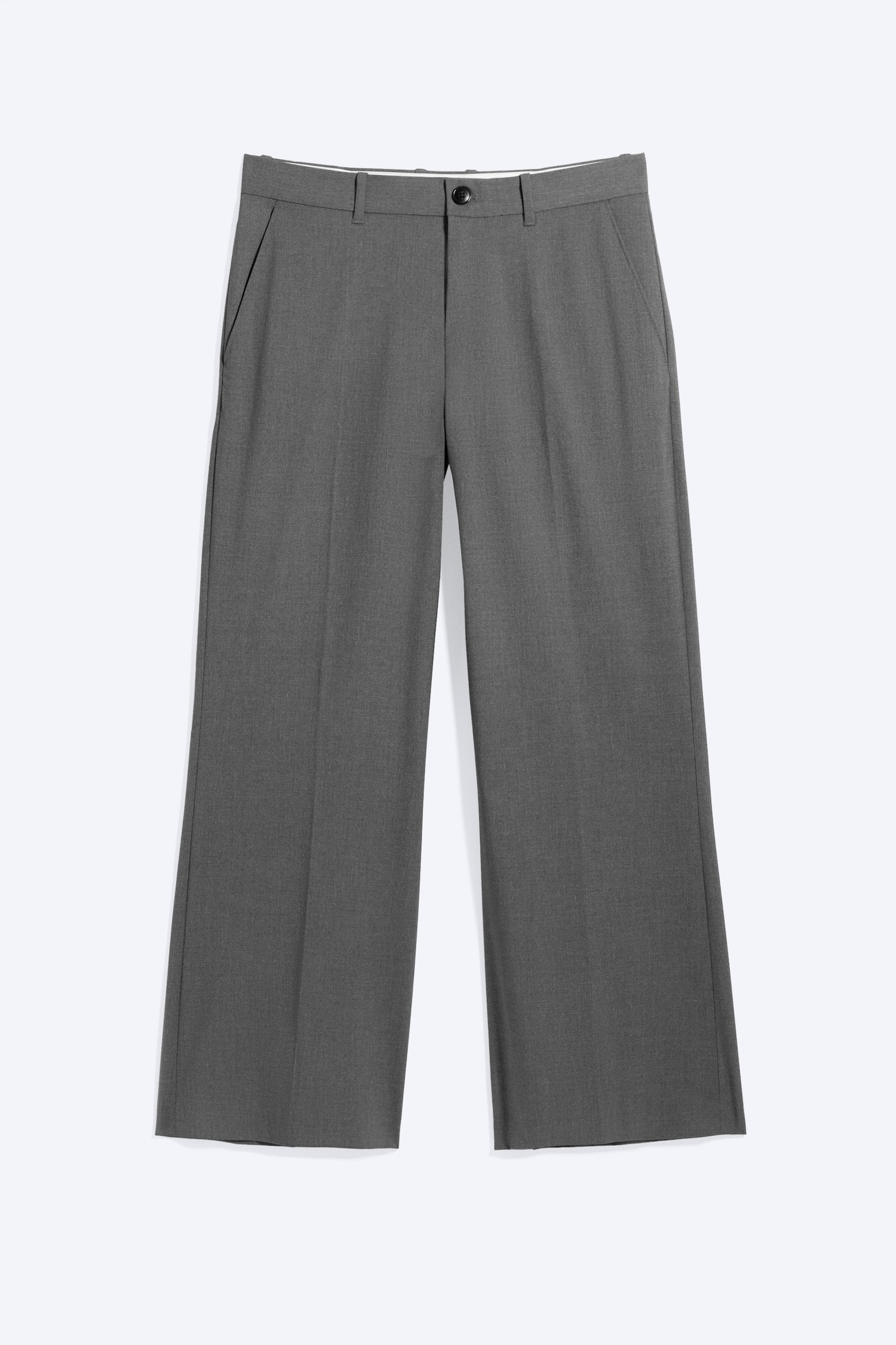 Un paio di pantaloni grigi a vita bassa da abito è mostrato frontalmente, presentando una vestibilità rilassata con una leggera gamba a zampa. I pantaloni sono caratterizzati da una chiusura con zip e bottone nero, tasche laterali oblique e pieghe stirate che scendono lungo il davanti di ciascuna gamba.