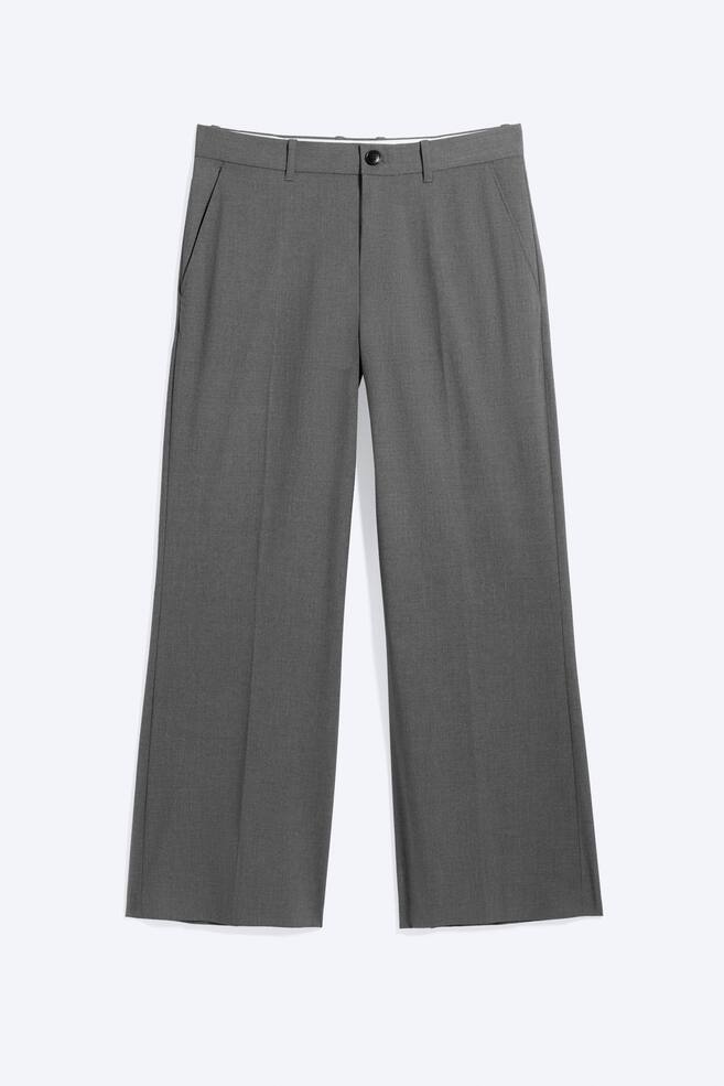 Un par de pantalones de traje de talle bajo en color gris se muestra desde el frente, exhibiendo un ajuste relajado con un sutil corte de bota. Los pantalones presentan una bragueta con cremallera y cierre de botón negro, bolsillos laterales inclinados y pliegues marcados que recorren la parte delantera de cada pierna. - 1