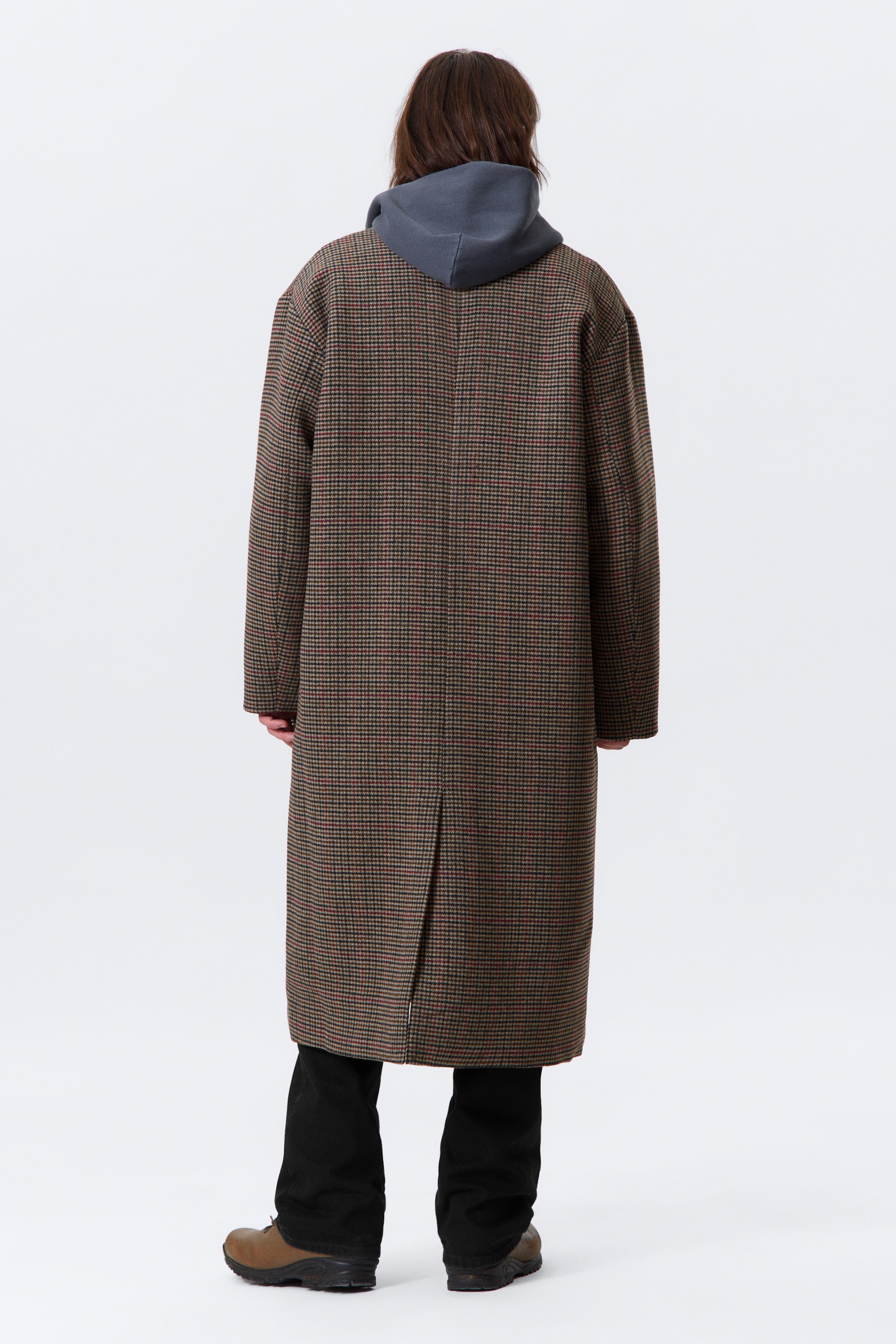Visualizza immagine più grande: Un lungo cappotto a micro quadri marrone e nero con sottili accenti rossi viene presentato dal retro, caratterizzato da una vestibilità rilassata e un unico spacco posteriore che si estende dall'orlo. Una felpa con cappuccio grigia viene indossata sotto, con il cappuccio che poggia sul colletto del cappotto.