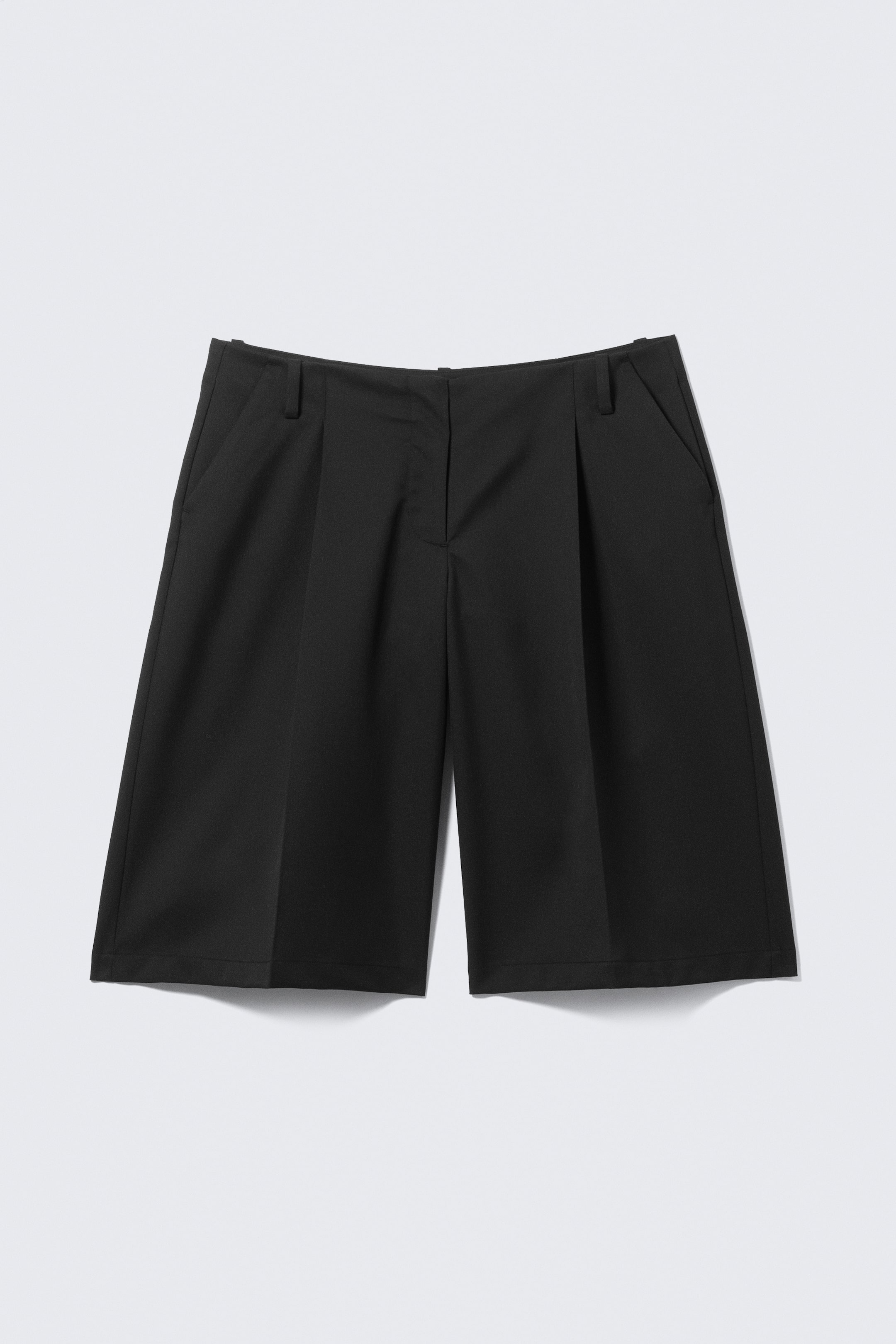 Visa större bild: Svarta shorts med vid passform och knälångt snitt, sedd framifrån. De har en mittframknäppning, hällor för bälte, djupa veck framtill och sidofickor.