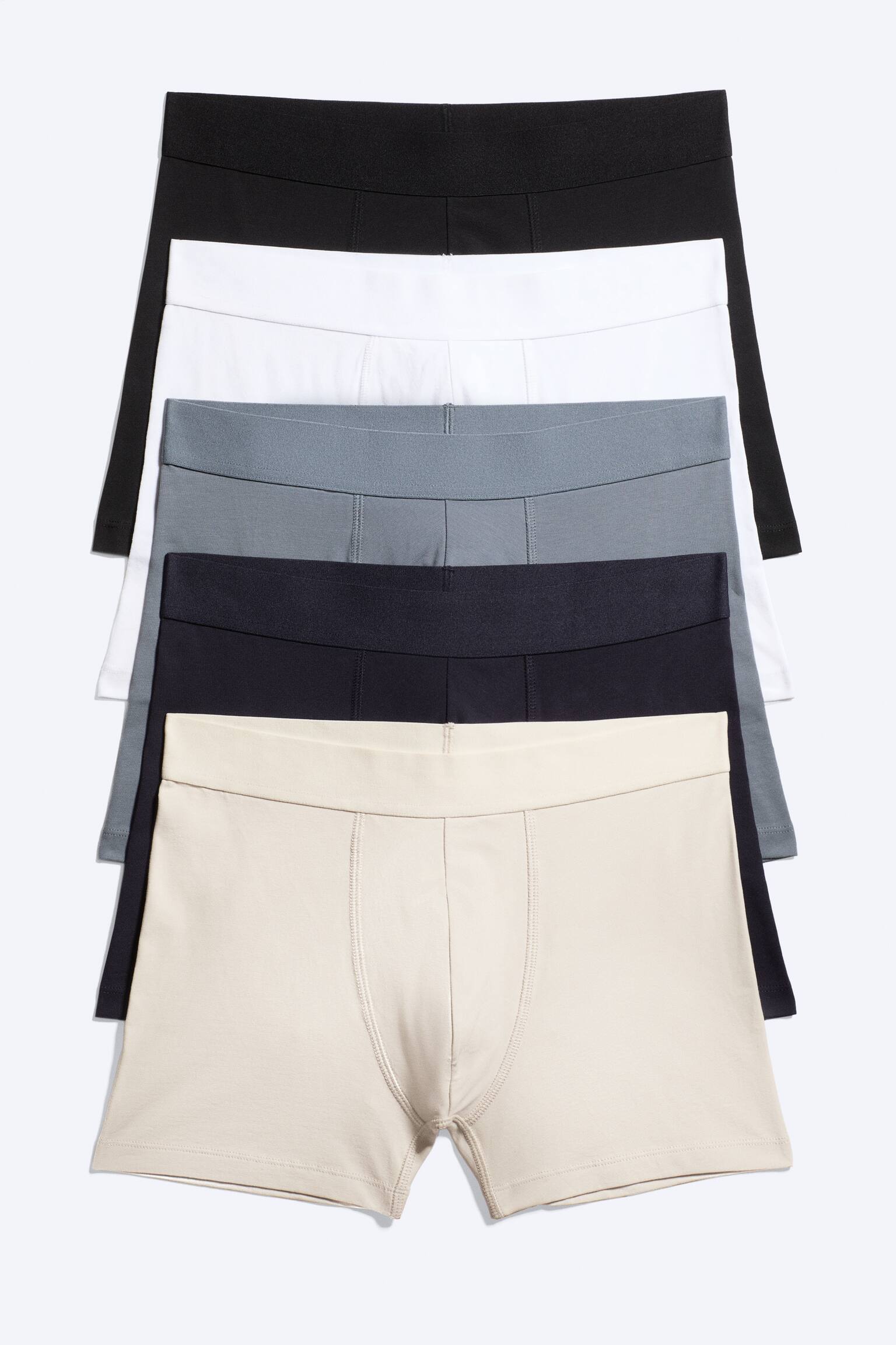 Cinque boxer sono disposti in pila, presentati frontalmente. Mostrano una sequenza di colori dall'alto verso il basso: nero, bianco, grigio, blu navy e beige. Ogni boxer presenta un'ampia fascia elastica in vita, e il paio beige in primo piano mostra una tasca sagomata con cuciture visibili.