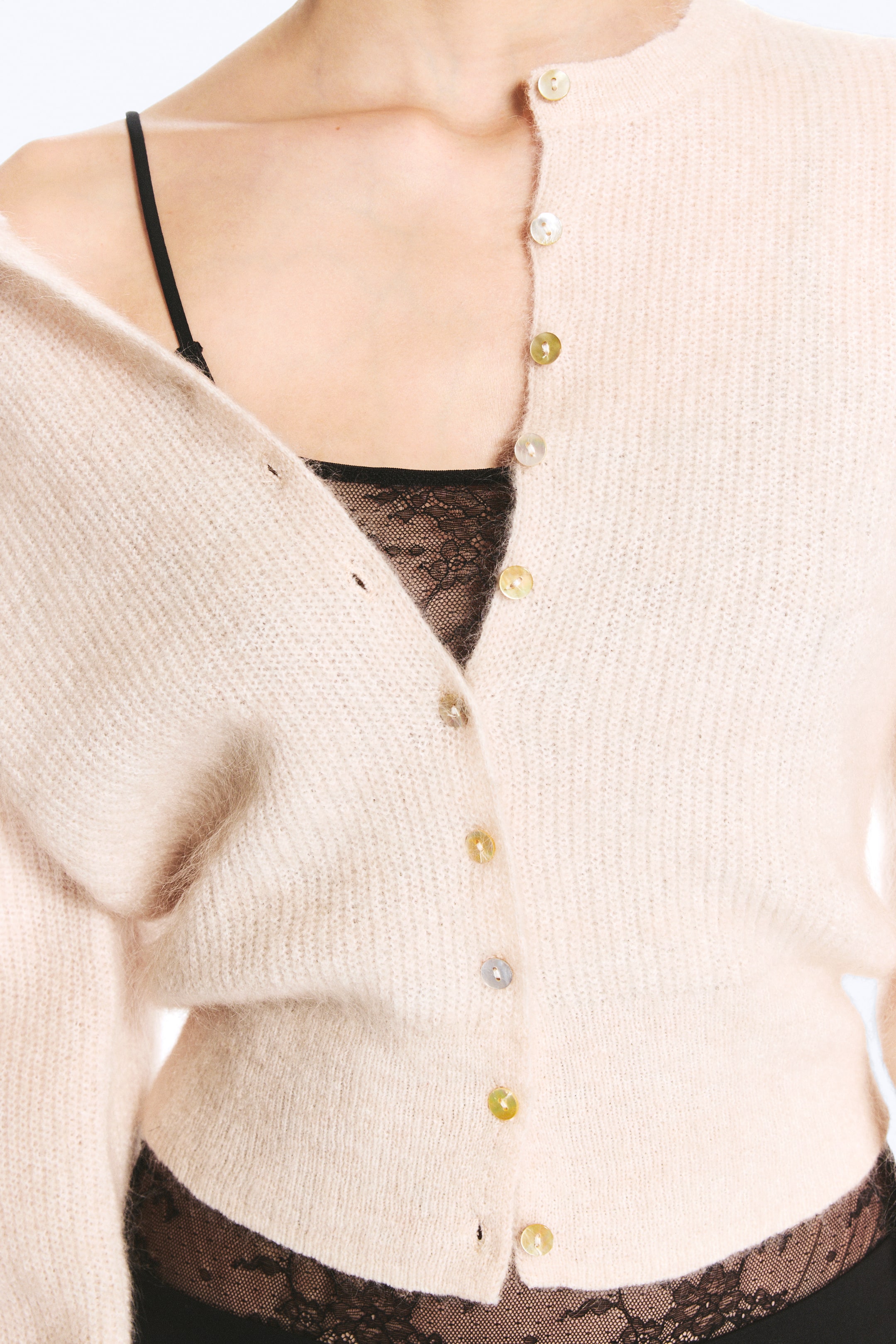 Agrandir l'image: Un cardigan en mélange de mohair, de teinte beige clair et en maille brossée, est porté par le mannequin, avec un côté drapé sur l’épaule. Le cardigan est doté de petits boutons irisés sur le devant, partiellement déboutonnés pour laisser apparaître un caraco en dentelle noire porté en superposition en dessous.