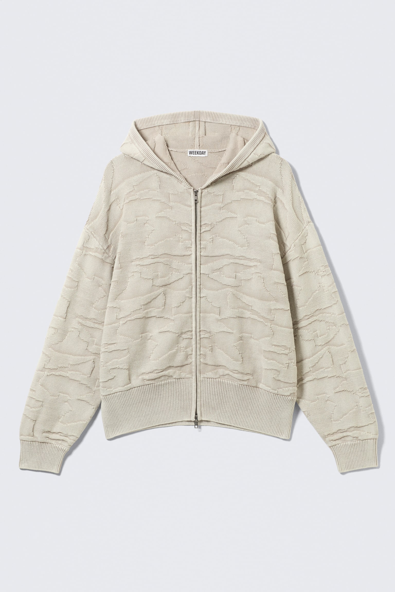 Een licht beige cardigan met kap toont zijn voorkant, met een getextureerd, abstract patroon over het lichaam en de mouwen. Het bevat een ritssluiting over de volledige lengte, geribde manchetten, een geribde zoom en een "WEEKDAY" merklint aan de binnenkant van de halslijn.