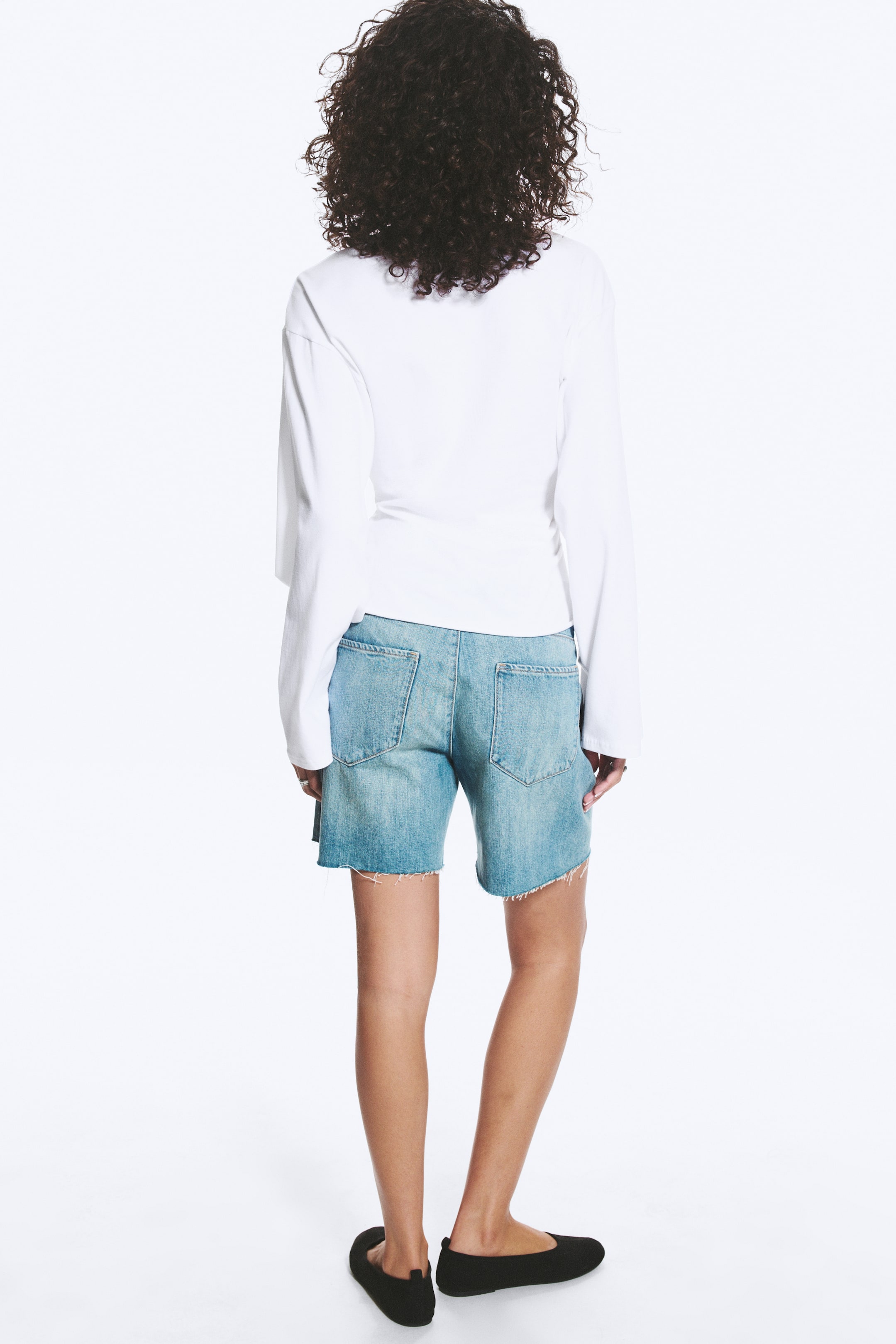 Grotere afbeelding bekijken: Een wit top met lange mouwen wordt van achteren gedragen, met een ontspannen pasvorm en gelaagd over licht gewassen denim shorts. De shorts hebben een ruwe zoom en twee zichtbare achterzakken, terwijl zwarte ballerina's de look compleet maken.