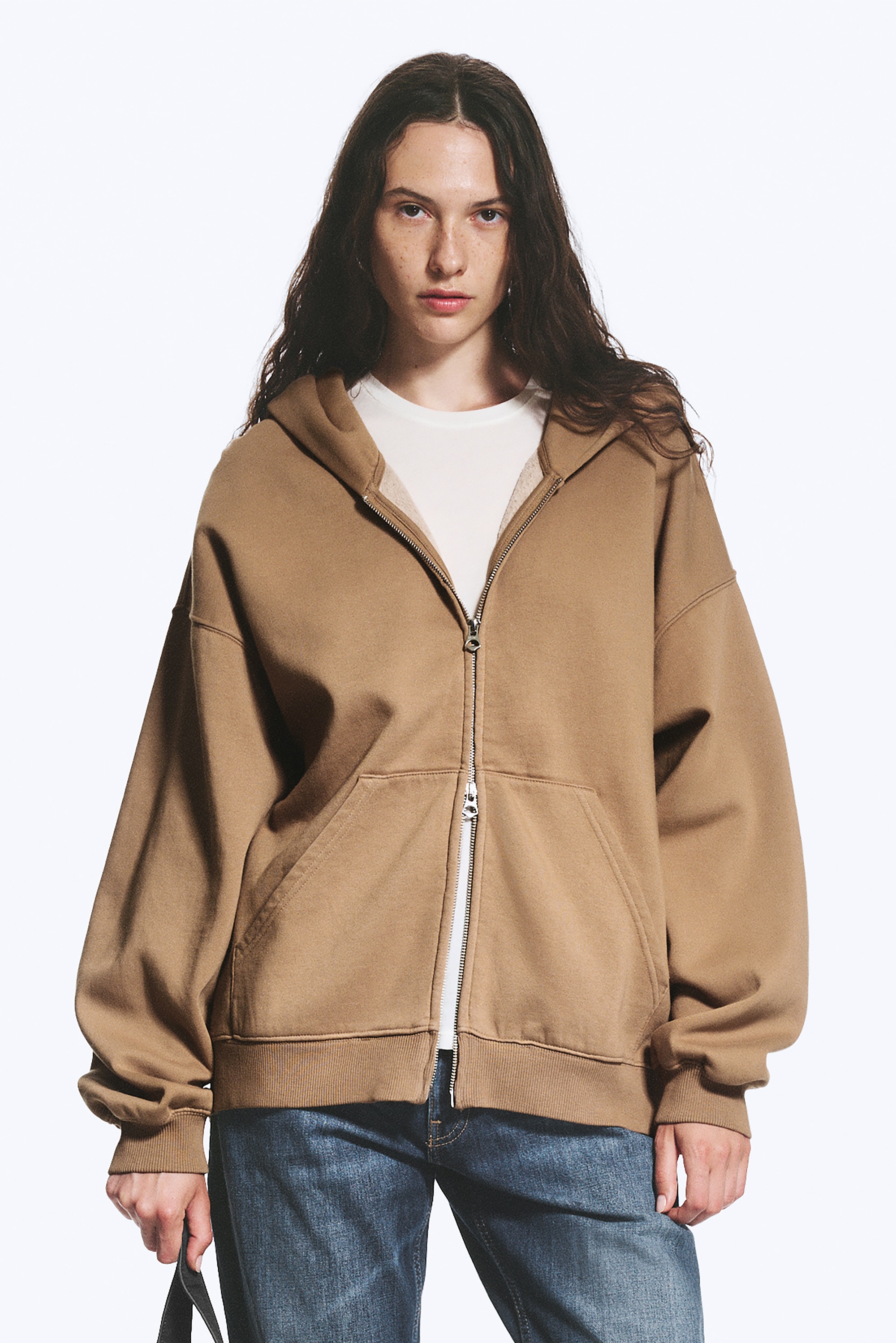 Un hoodie zippé marron est présenté de face, arborant une silhouette ample et décontractée. Il dispose d'une capuche spacieuse à double épaisseur sans cordons de serrage, d'épaules tombantes, d'une fermeture éclair cachée à double sens, de grandes poches, et de poignets et ourlet côtelés.