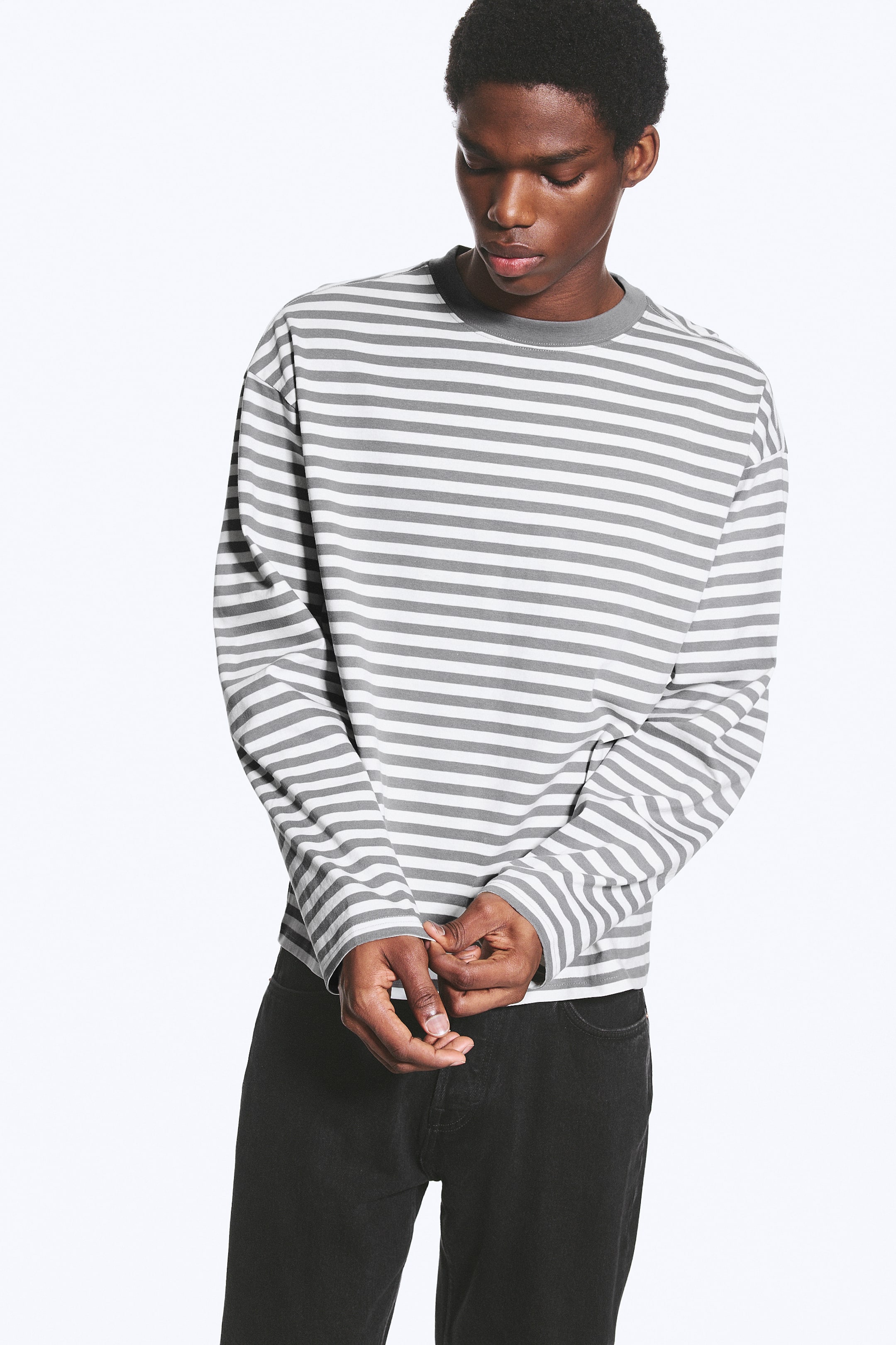 Grotere afbeelding bekijken: Een boxy T-shirt met lange mouwen en grijze en witte horizontale strepen wordt gedragen door het model. Het heeft een ronde hals met een grijze kraag en een losse pasvorm. Het model staat met de handen gevouwen, waardoor de rechte zoom van het shirt zichtbaar is.