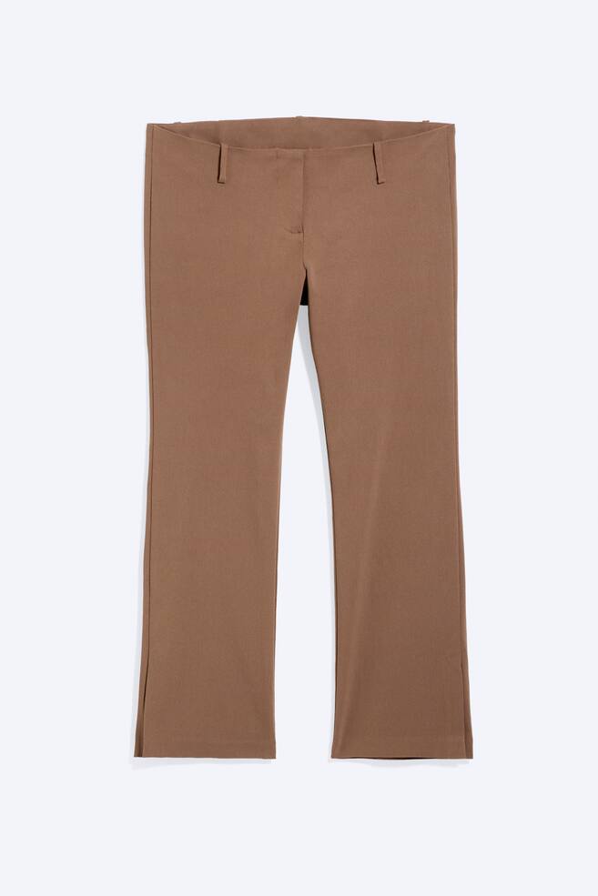 Los pantalones marrones presentan un frente plano con dos trabillas visibles para el cinturón y una discreta costura central. Las piernas tienen un ligero acampanado hacia el dobladillo, cada una con una pequeña abertura en la costura exterior. - 1