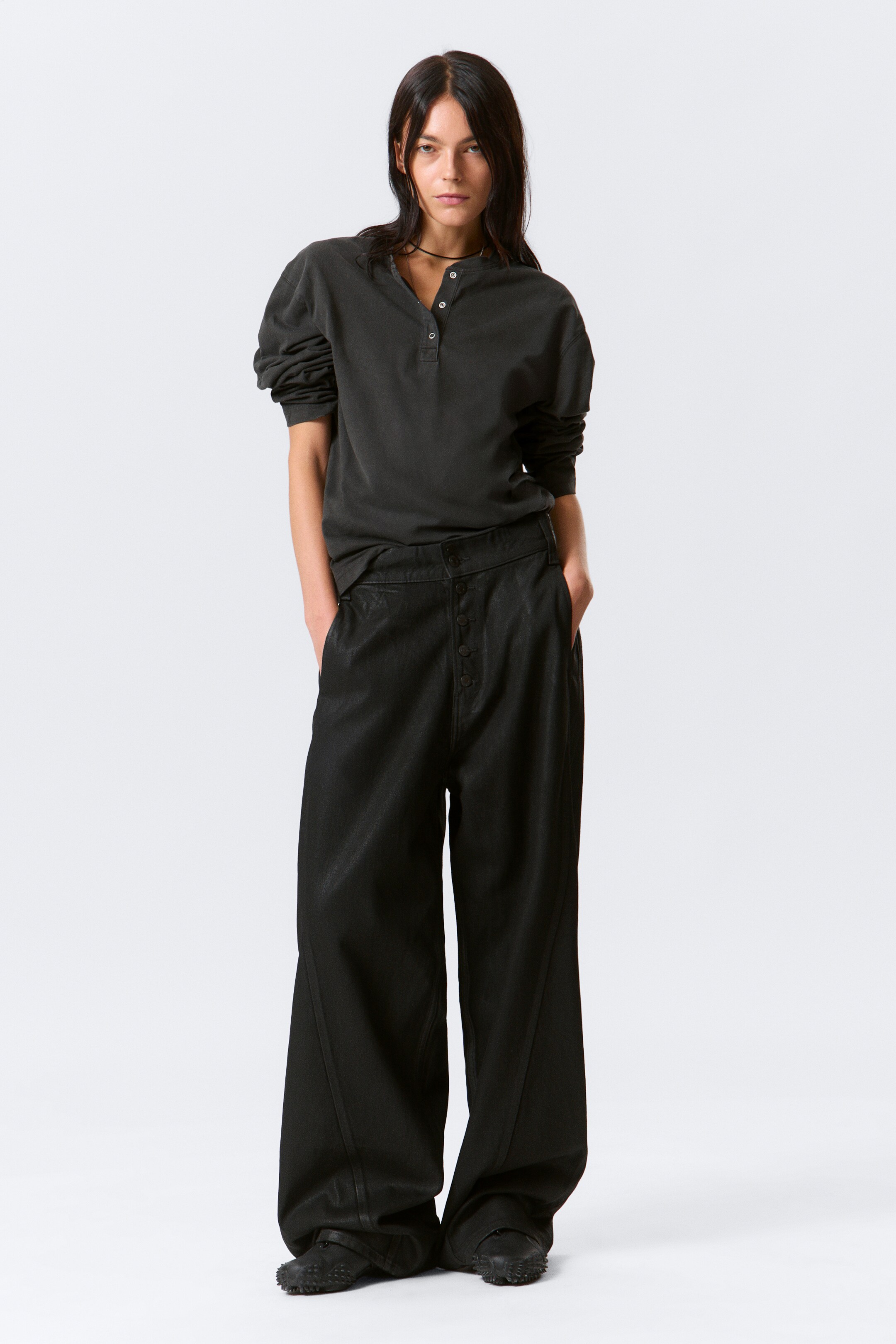 View larger image: A dark charcoal henley shirt is being presented, featuring a four-button placket and voluminous long sleeves. It is being paired with black wide-leg pants, which are showcasing a prominent button fly closure and vertical seams running down the front of each leg.