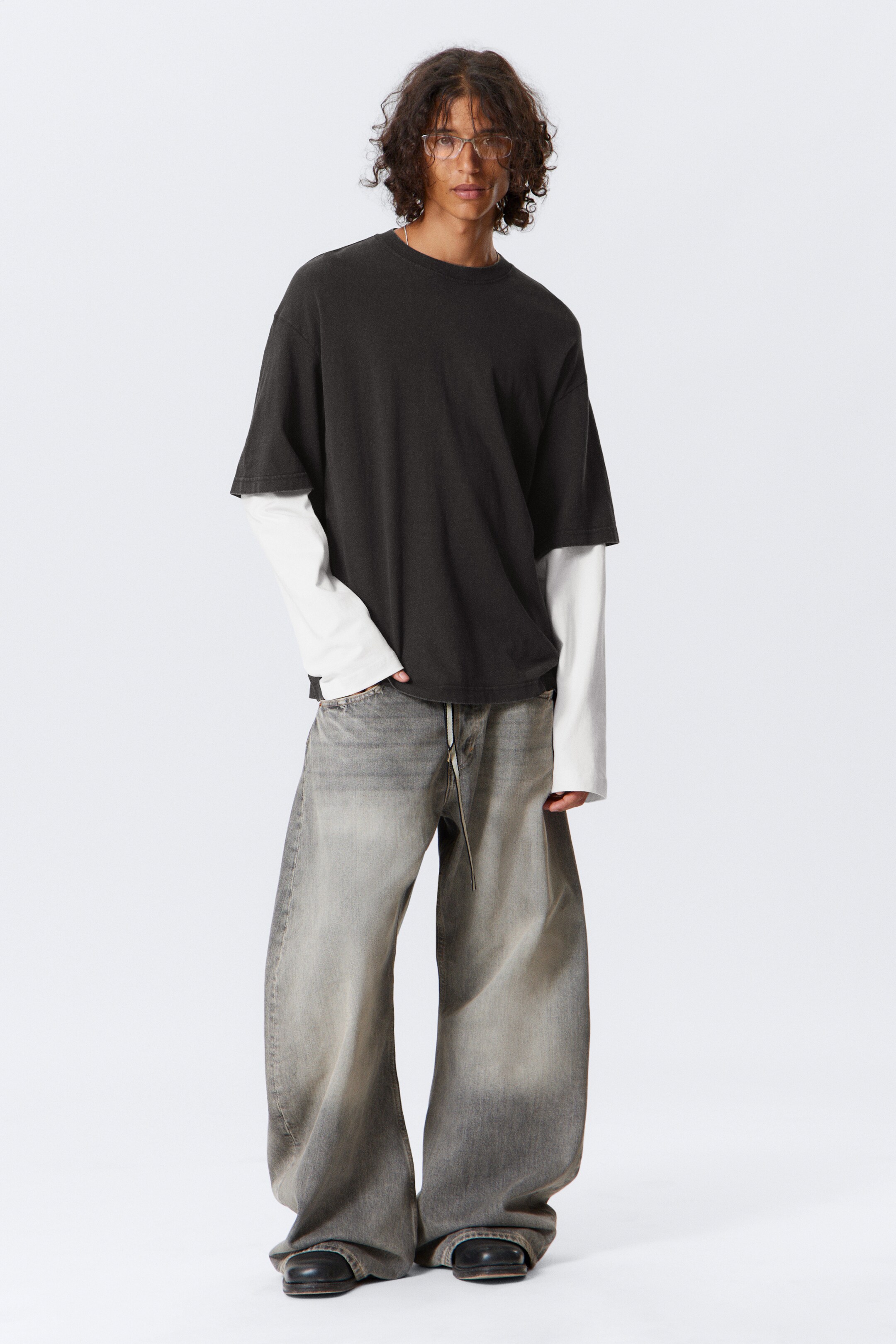 View larger image: An oversized, faded black short-sleeved t-shirt is being worn over a white long-sleeved t-shirt, creating a layered look. Very wide-leg, faded grey jeans with a visible drawstring are being worn, their hems pooling over dark shoes.