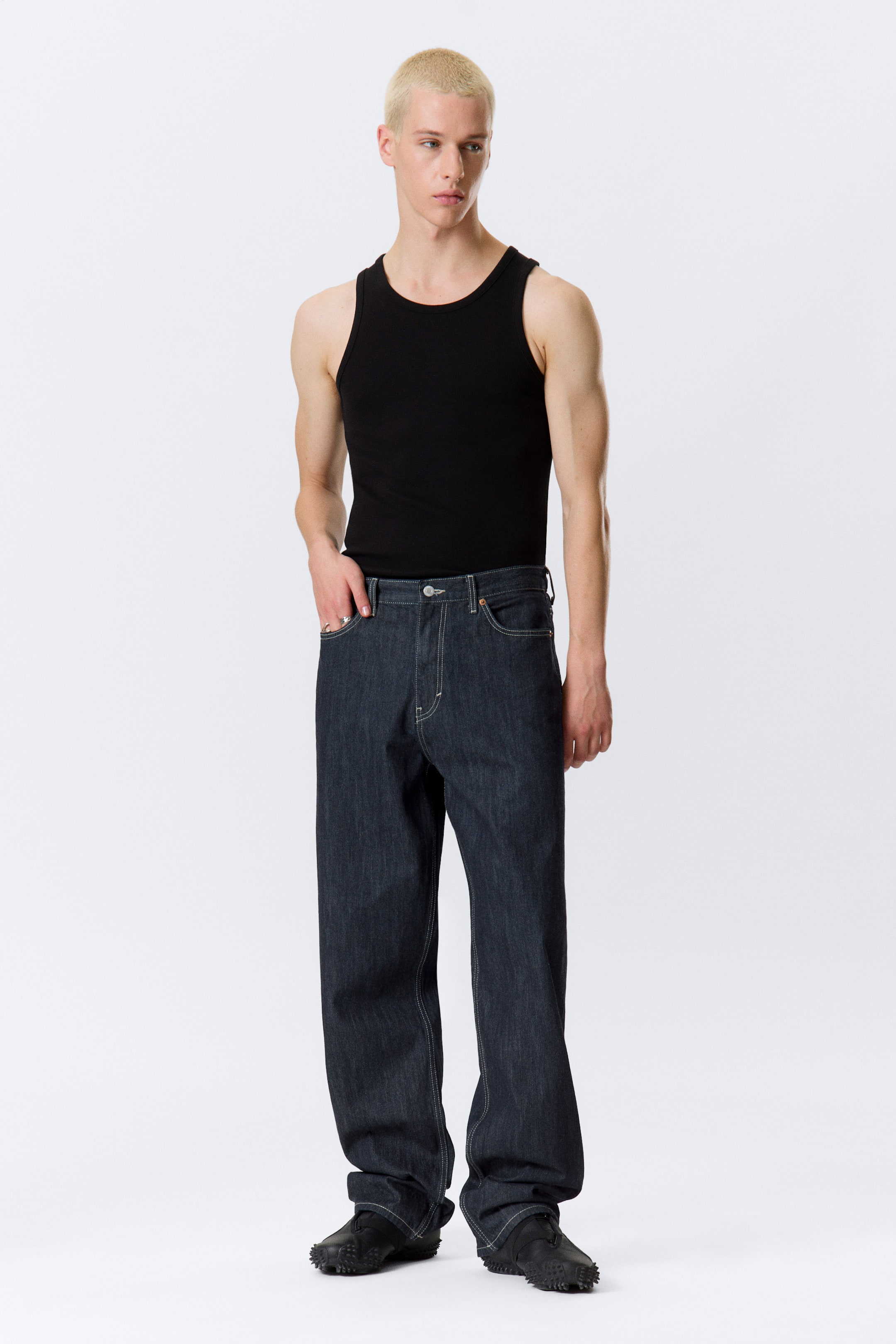 Grotere afbeelding bekijken: Losse, rechte jeans in een donkere wassing met een lange lengte en contrasterende stiksels worden gedragen door het model. Deze jeans hebben een klassiek 5-pocket ontwerp en de hand van het model rust in een van de voorzakken.