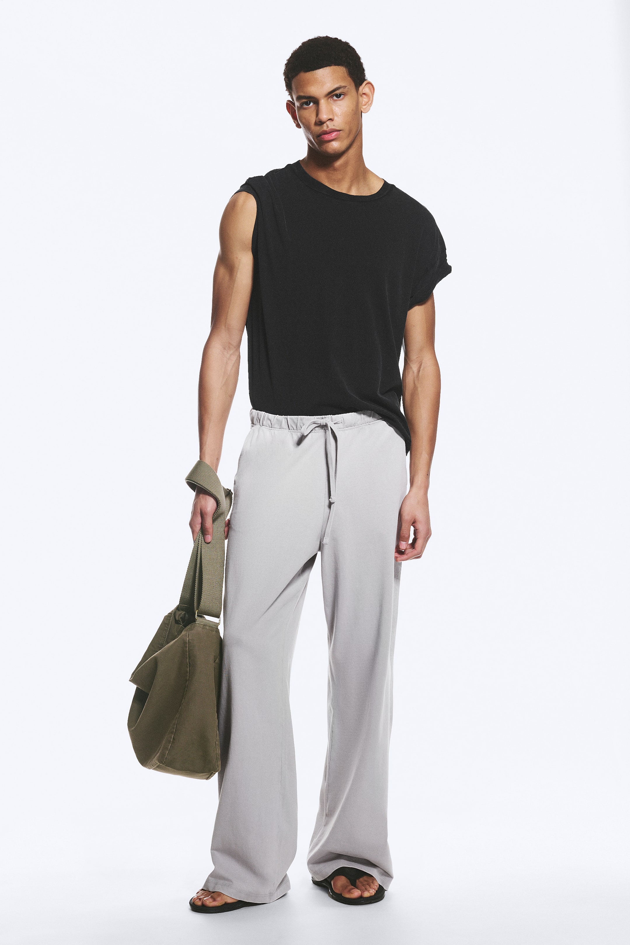 Se større billede: Lysegrå bukser med brede ben, der har en snøre i taljen og en afslappet pasform, stylet med en sort ærmeløs crew-neck top, en olivengrøn messenger-taske i hånden og sorte klipklapper.