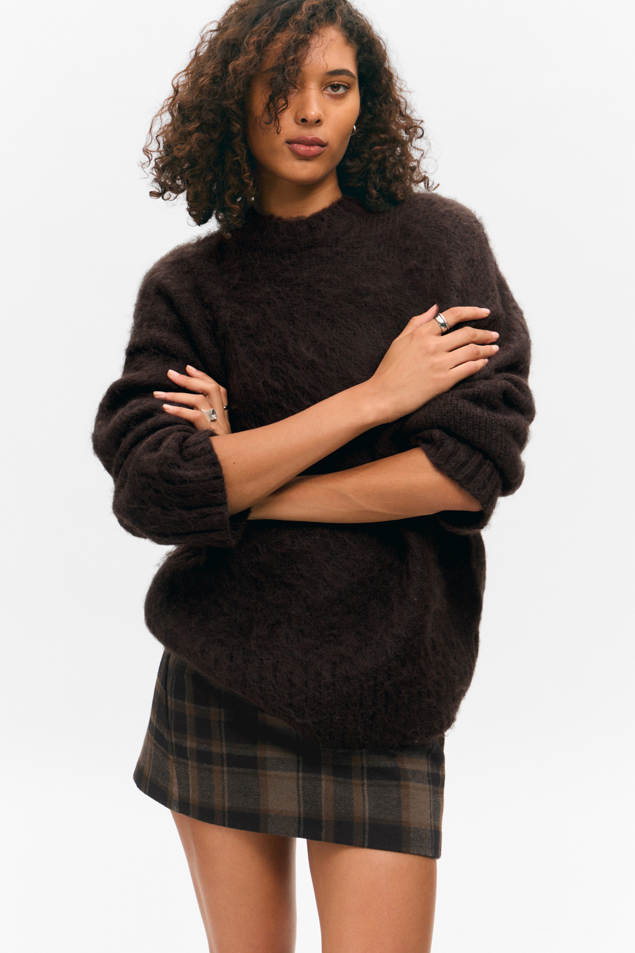 Ingrandisci l'immagine: Il maglione marrone scuro presenta una texture pelosa, maglia spazzolata e uno scollo rotondo. È presentato di fronte, mostrando la sua vestibilità ampia e le maniche lunghe con polsini a costine, mentre l'orlo è appoggiato su una mini gonna a quadri marrone e verde.