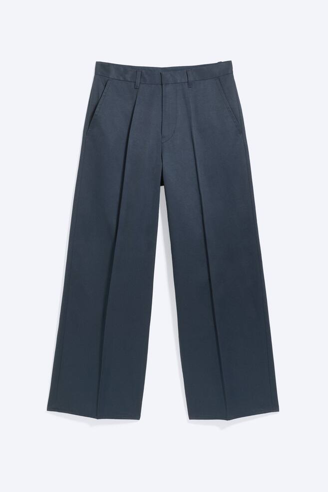 Un par de pantalones de sarga azul oscuro de corte holgado se presenta de frente, con pliegues prensados en la parte delantera de cada pierna, bolsillos laterales inclinados y bragueta con cremallera, cinturilla y trabillas para el cinturón. - 1