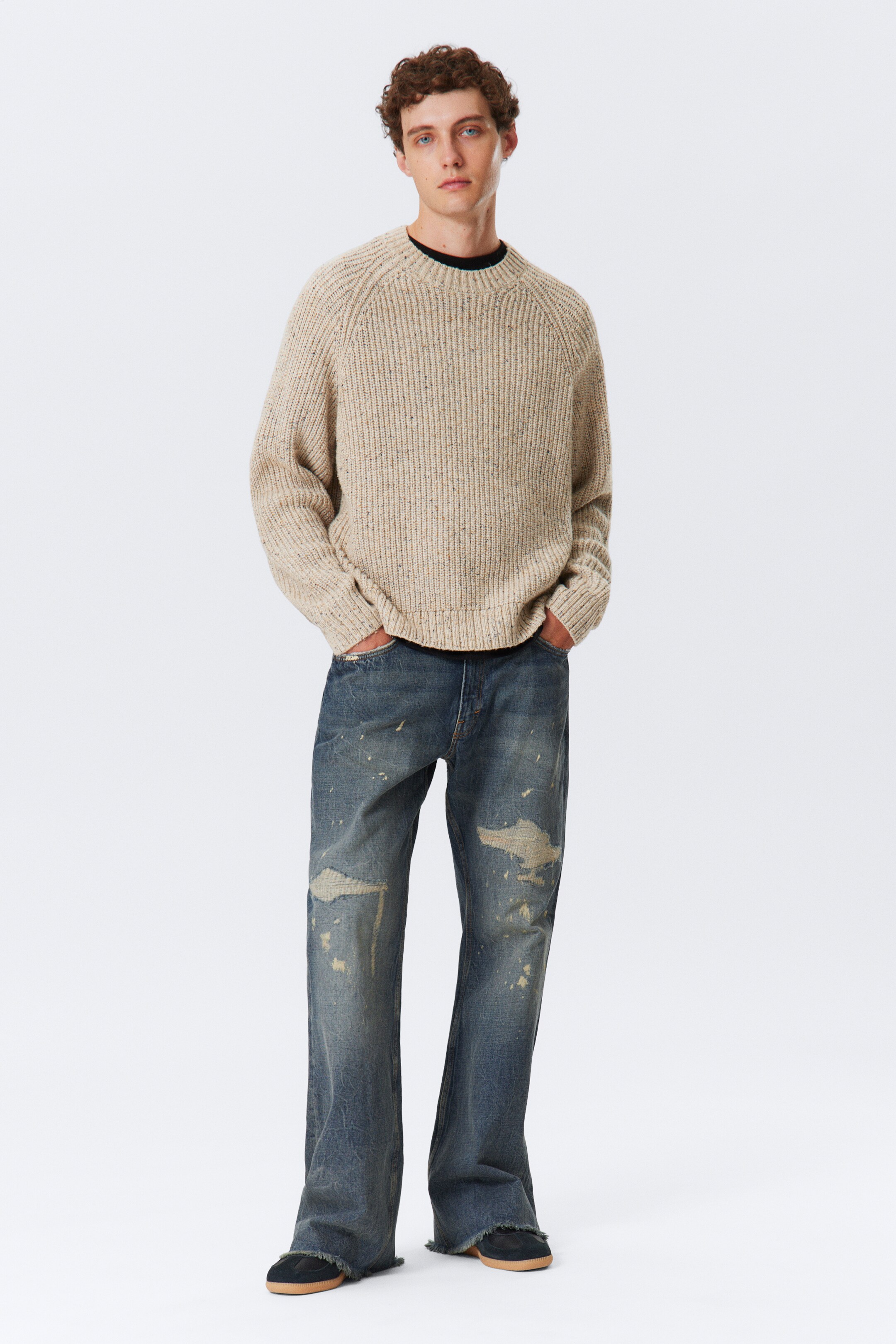 Agrandir l'image: Un pull en tricot côtelé beige clair, moucheté, avec des manches raglan et un sous-vêtement foncé est porté, assorti à un jean en denim bleu à jambes larges et usé, avec des zones délavées et un ourlet effiloché tombant sur des chaussures foncées.
