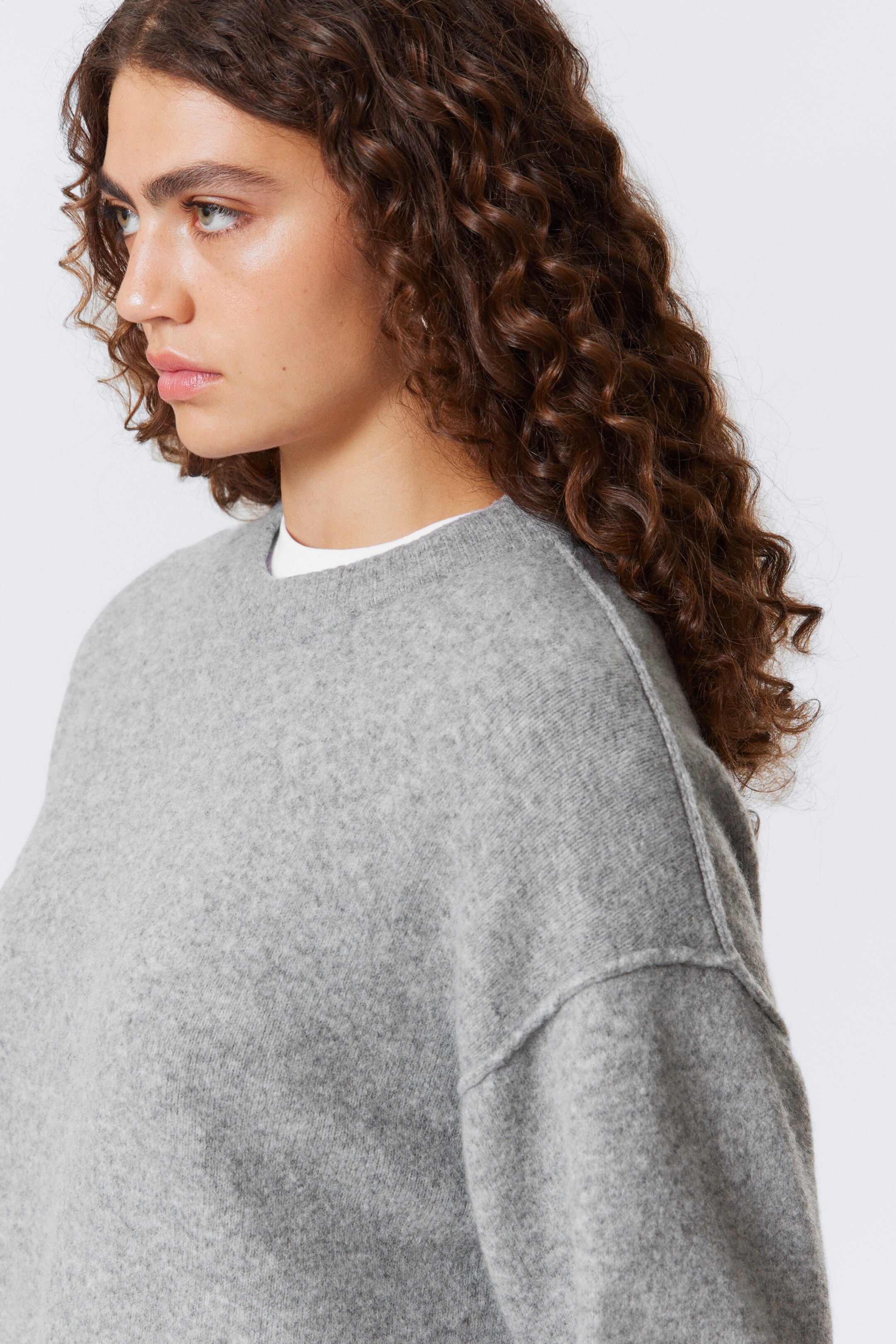 Se større billede: En lysegrå sweater med rund hals præsenteres fra en sidevinkel, med en afslappet pasform og sænkede skuldre med synlige, eksponerede sømme, der løber ned ad ærmerne. En hvid t-shirt er lagdelt underneden, der titter frem ved halsudskæringen.