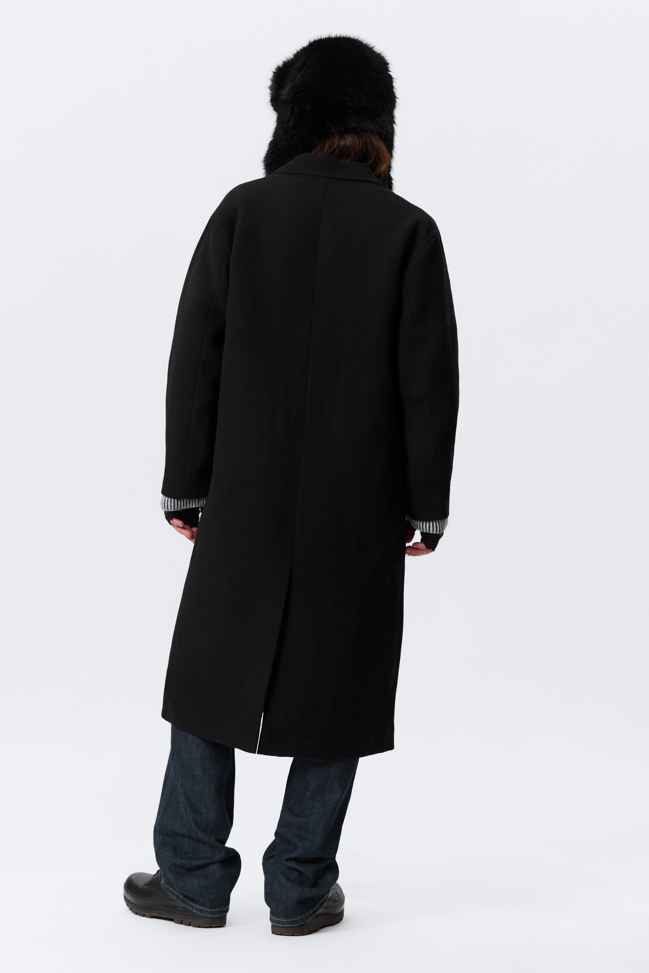 View larger image: A black long coat is being presented from the back, featuring a single back vent extending from the mid-thigh and revealing striped cuffs extending from under its long sleeves.