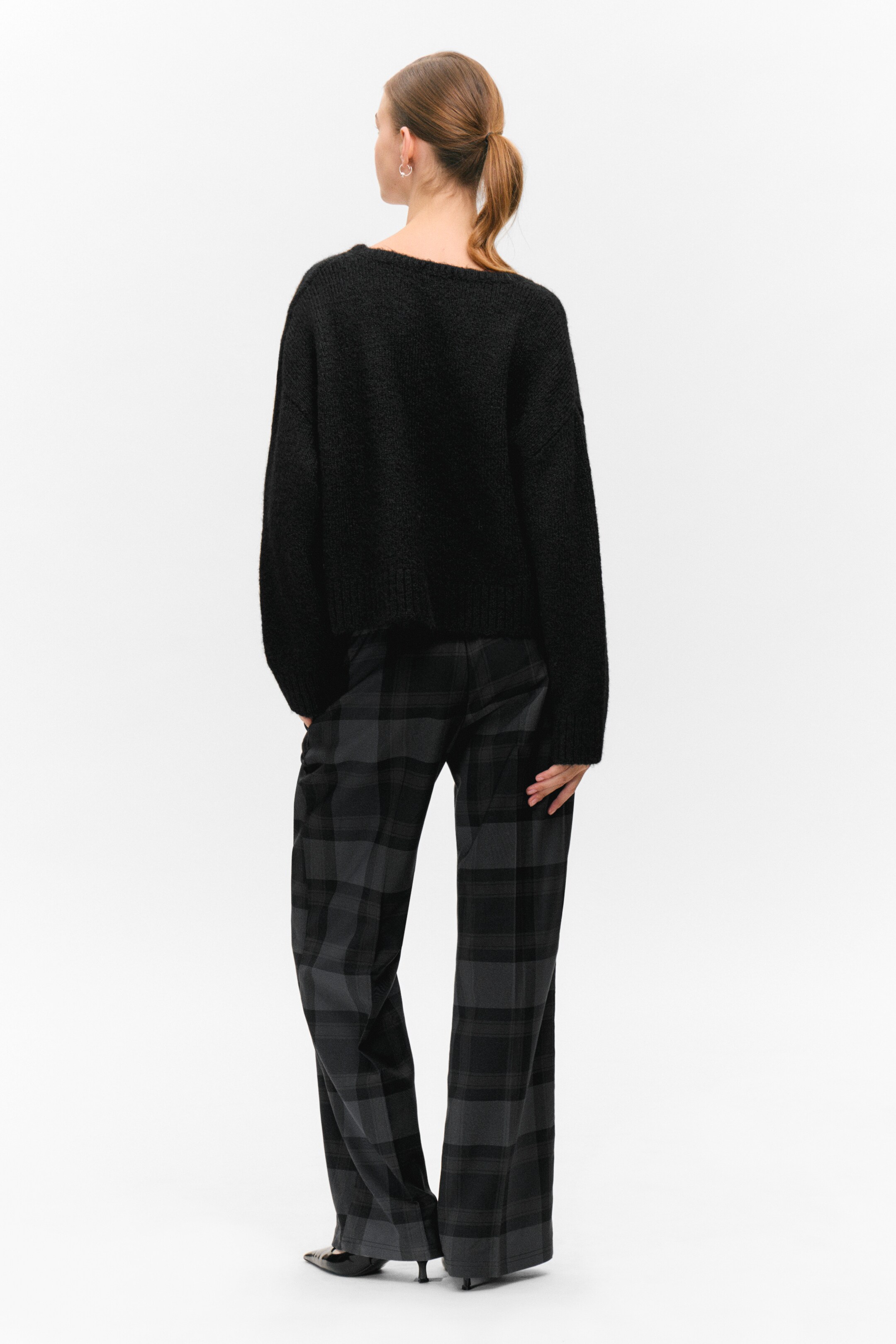 Visualizza immagine più grande: Un maglione nero a maglia pelosa con una vestibilità rilassata e corta viene presentato dal retro, mostrando il suo ampio scollo e le maniche lunghe. Viene abbinato a pantaloni a gamba larga con un motivo a quadri di colore antracite e nero.