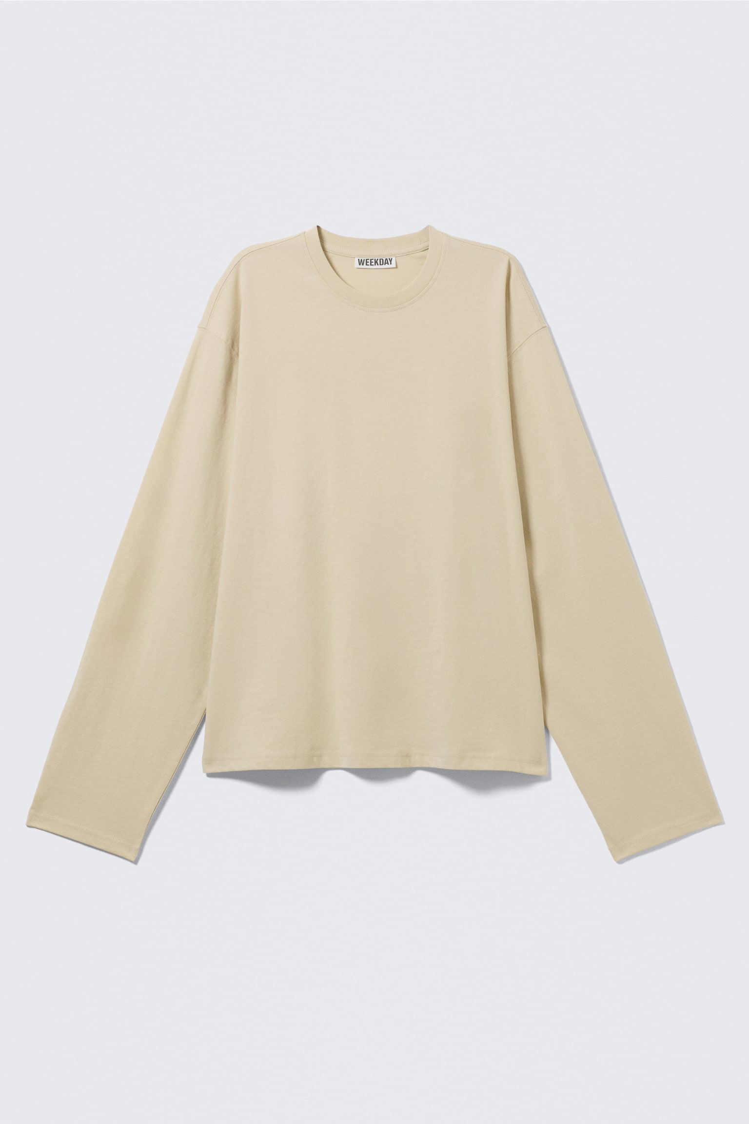 Un tricou bej cu mâneci lungi este prezentat din FAȚĂ, având o croială lejeră și boxy. Prezintă un guler rotund cu nervuri și o etichetă vizibilă „WEEKDAY”, precum și un tiv drept.