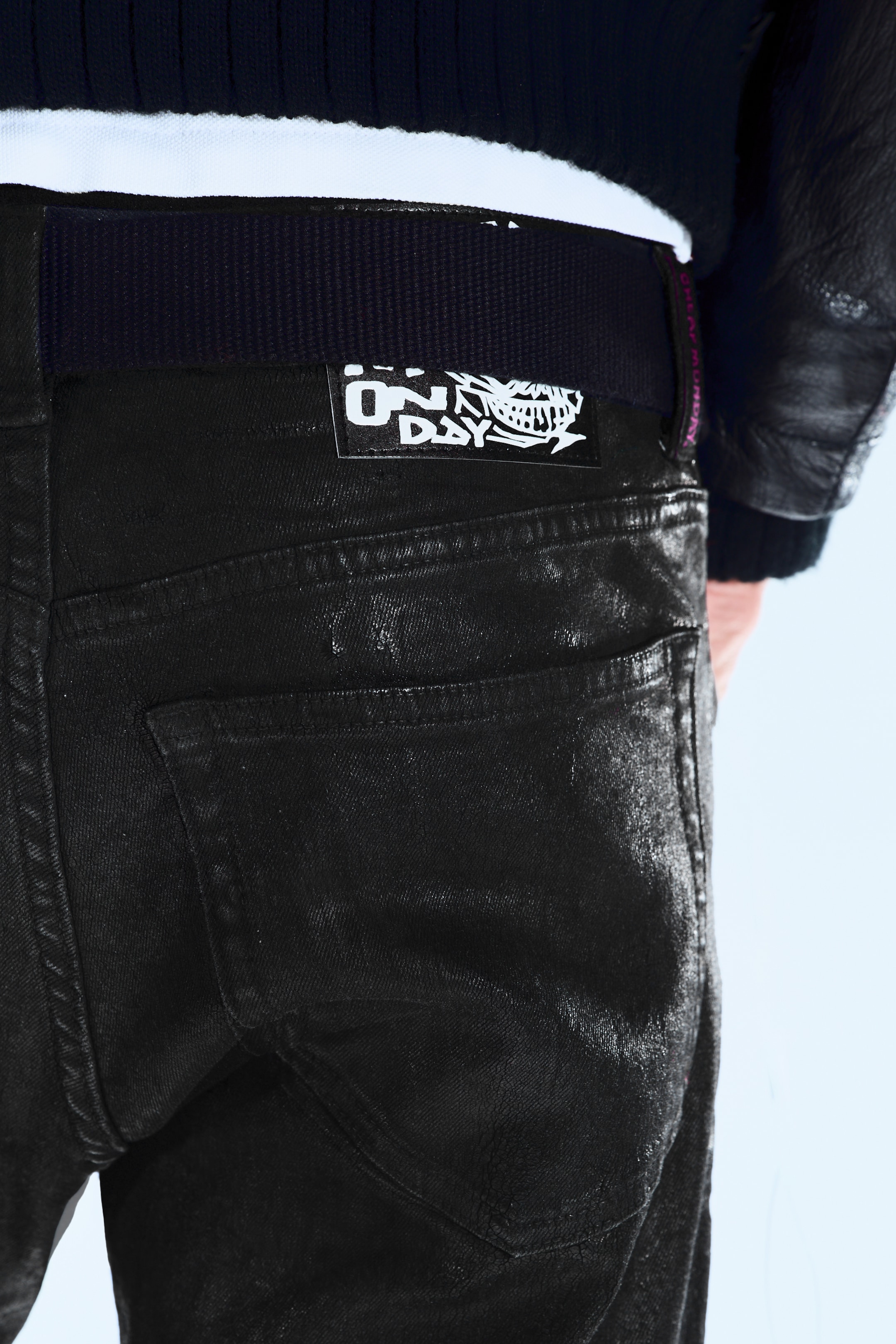 Visualizza immagine più grande: I jeans neri sono presentati da dietro e di lato, mostrando una finitura leggermente cerata o rivestita. Presentano due tasche posteriori applicate e una cintura a coste scura che mostra un'etichetta rettangolare bianca con testo nero e un grafico.