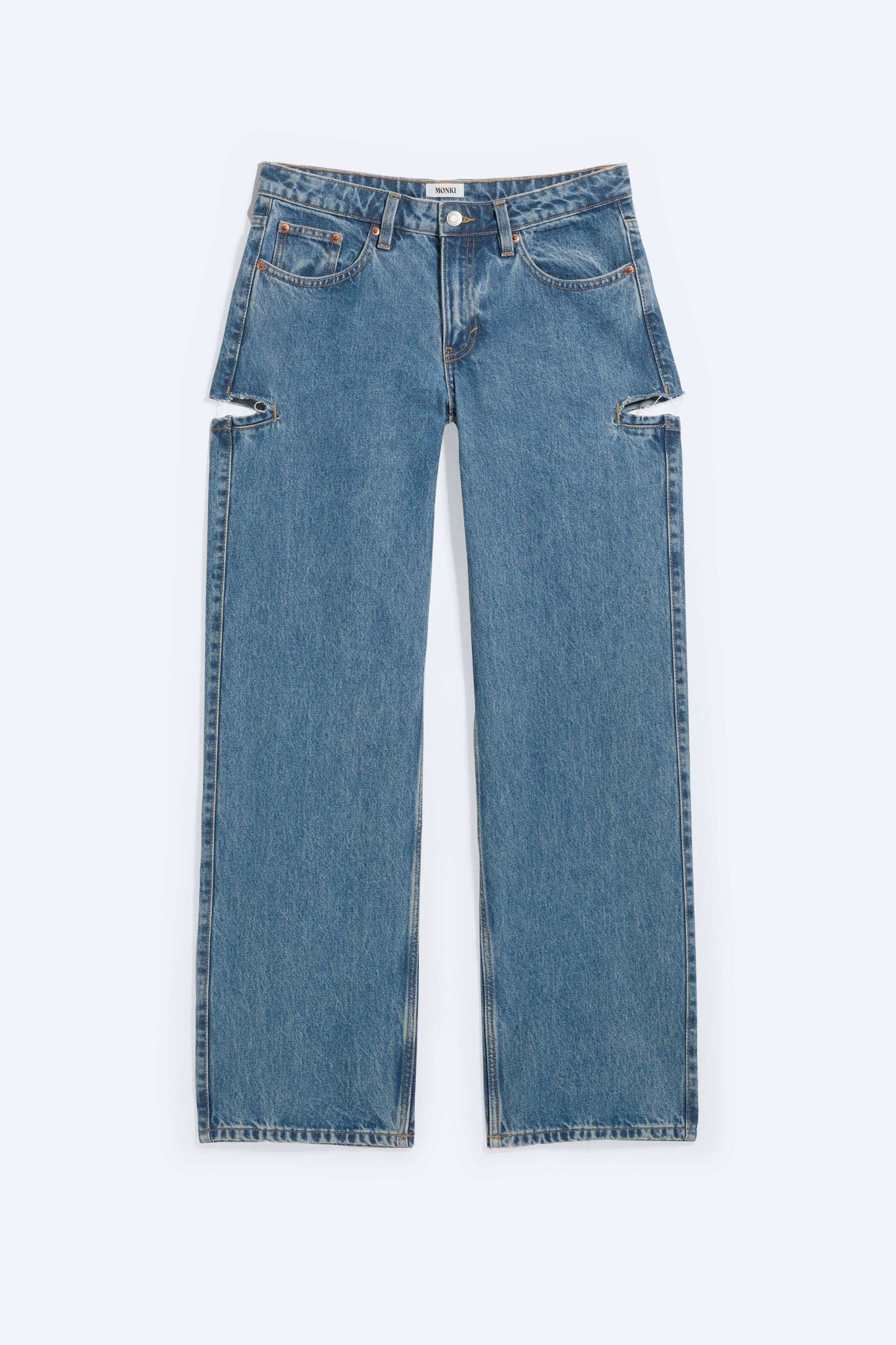 Agrandir l'image: Un jean bleu taille basse est présenté de face, avec une coupe ample et une jambe évasée. Il est conçu avec une construction classique à cinq poches, une fermeture éclair, et des découpes sur le haut des cuisses extérieures avec des bords effilochés visibles, créant un look déstructuré.