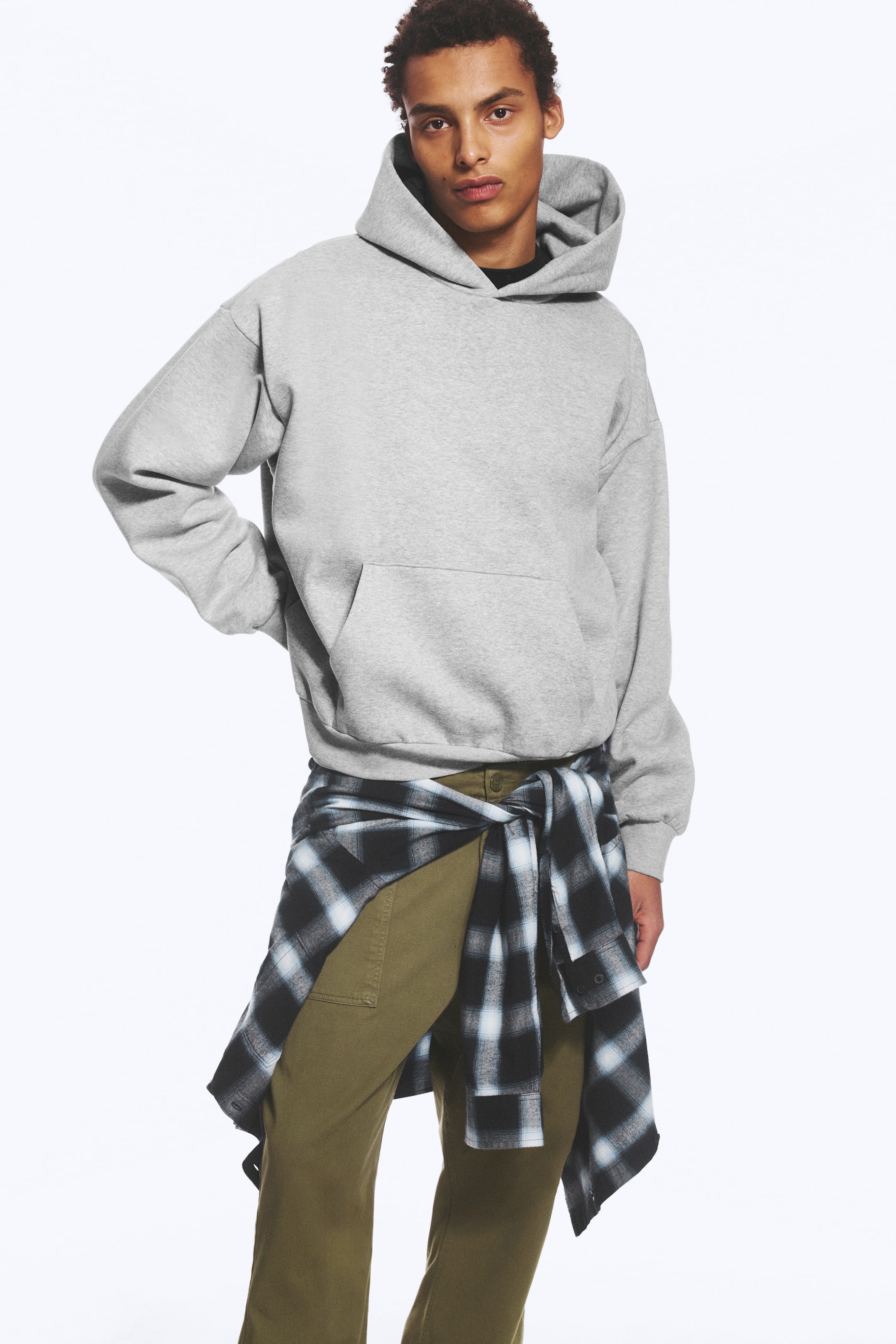 Vezi imaginea mai mare: Un hanorac gri din fleece de bumbac, cu croială lejeră, umeri căzuți și buzunar tip kangaroo, purtat de model. Modelul pozează cu o mână sprijinită pe șold, asortând hanoracul cu pantaloni verde măsliniu și o cămașă în carouri legată în jurul taliei.