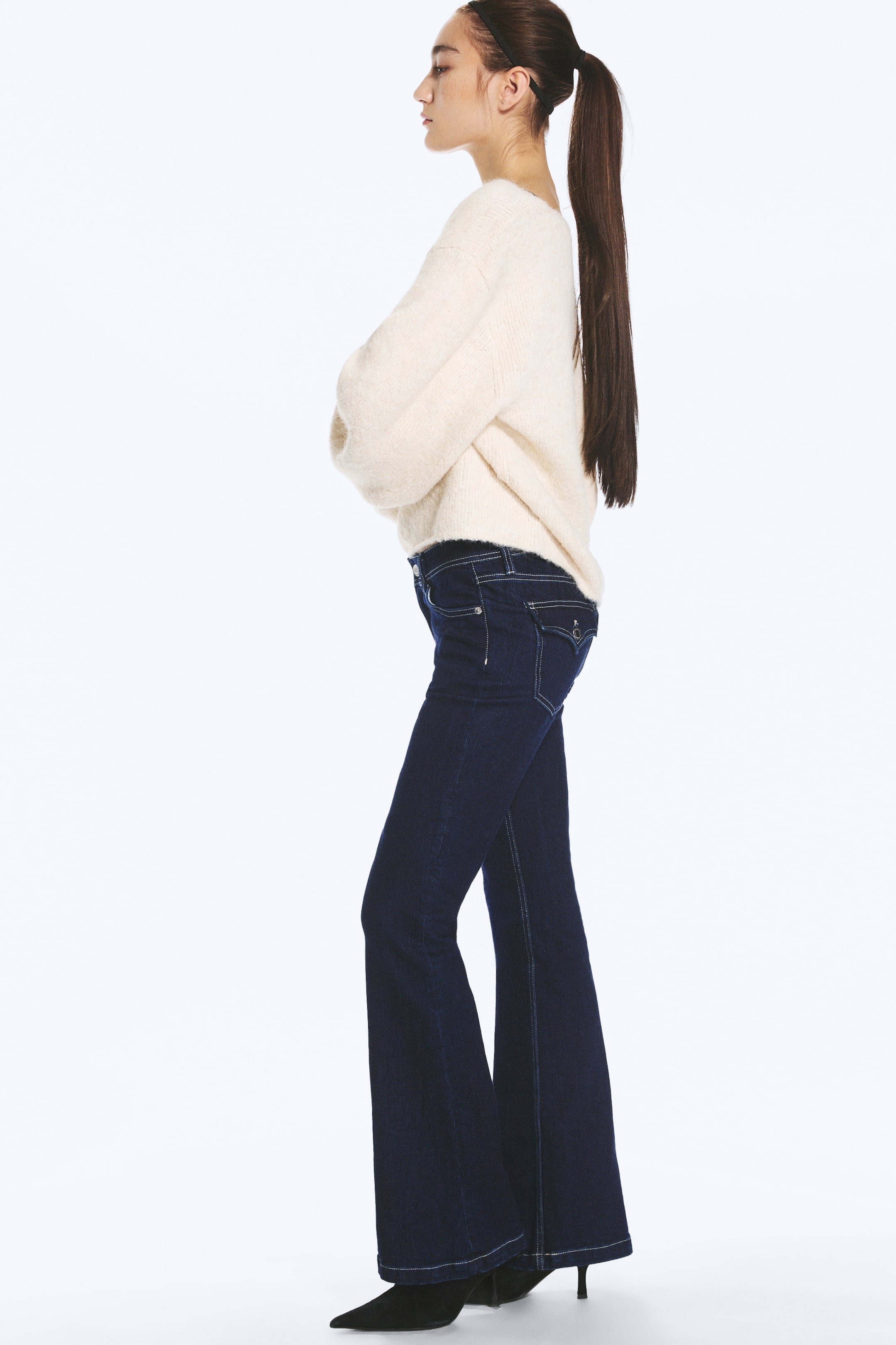 Ver imagem maior: Um par de jeans azul-escuro, de cintura baixa, com corte slim e perna evasé, é usado pela modelo, que está de perfil. Os jeans apresentam um design clássico de cinco bolsos, com um bolso traseiro com aba e botão, e estão combinados com uma camisola de malha cor de creme e sapatos pretos de salto alto com bico fino.