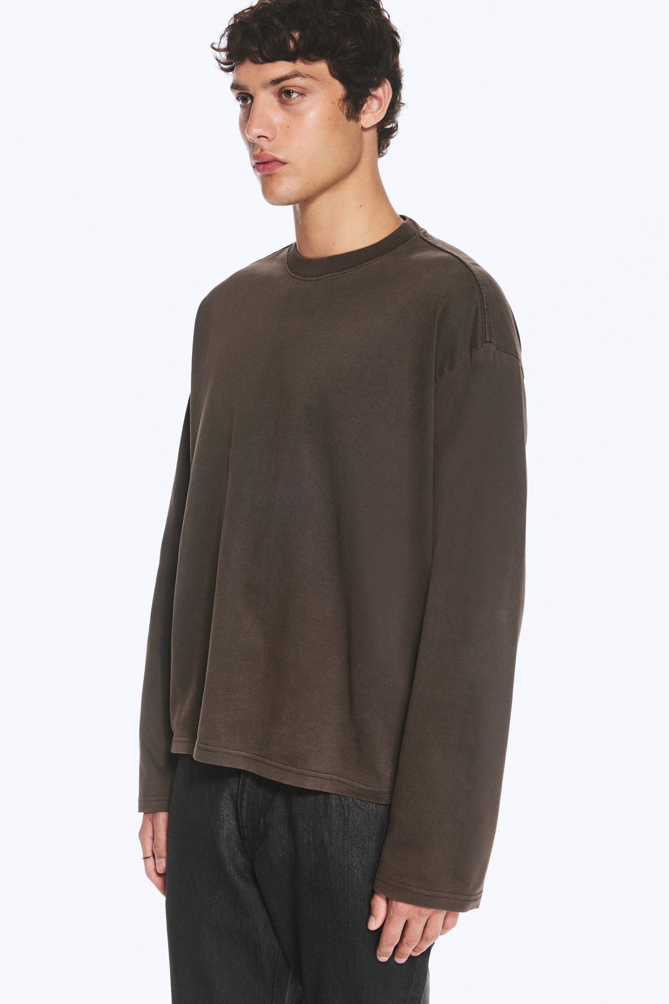 Visa större bild: En boxig långärmad T-shirt i mörkbrunt bärs av modellen, med rund halsringning, nedsänkta axlar och lös passform med rak fåll. T-shirten kombineras med mörka, strukturerade byxor.