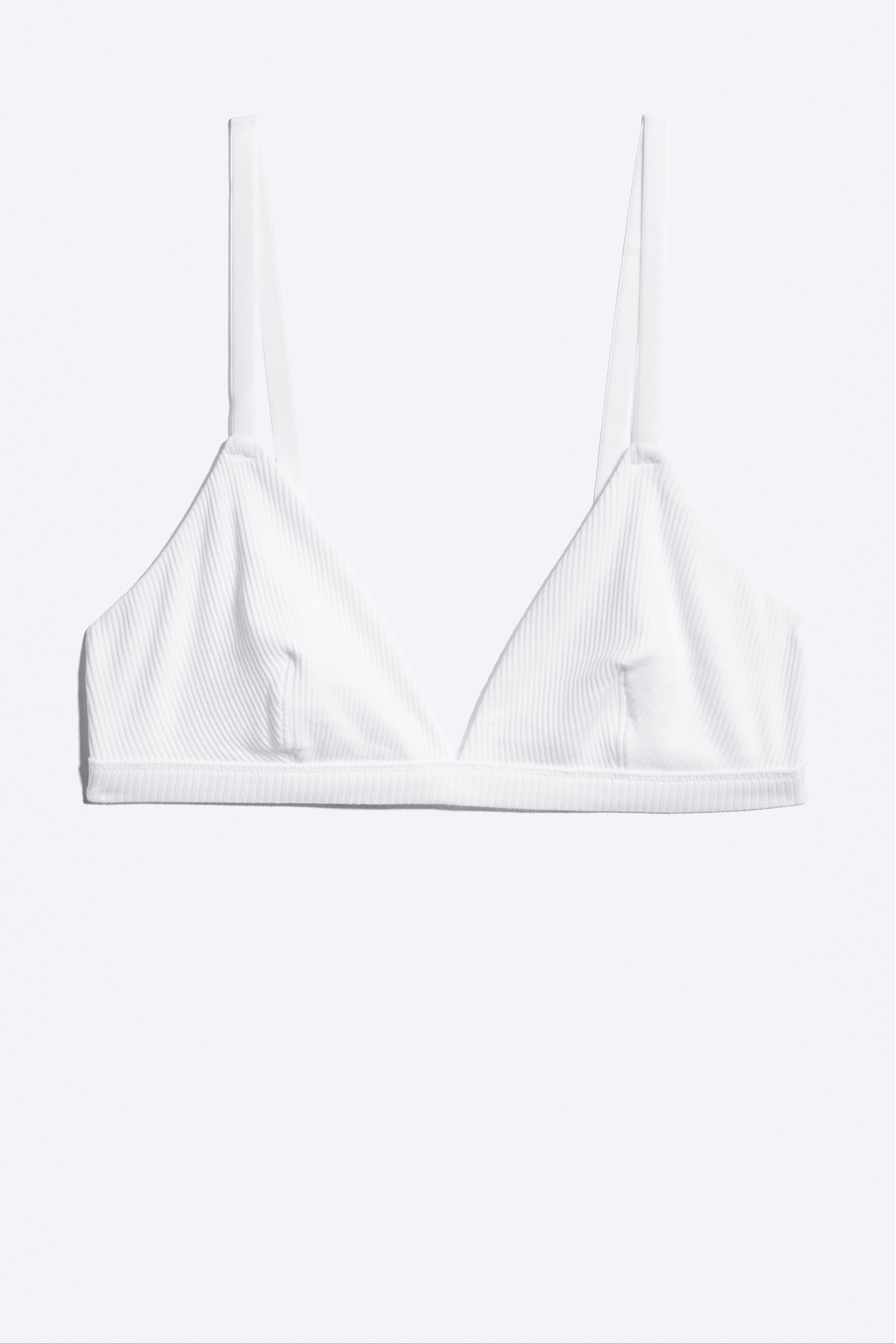 Un reggiseno a triangolo morbido bianco, a coste, con sottili spalline e un ampio sottoseno, indossato dalla modella. Il reggiseno è abbinato a slip bianchi a coste coordinati.