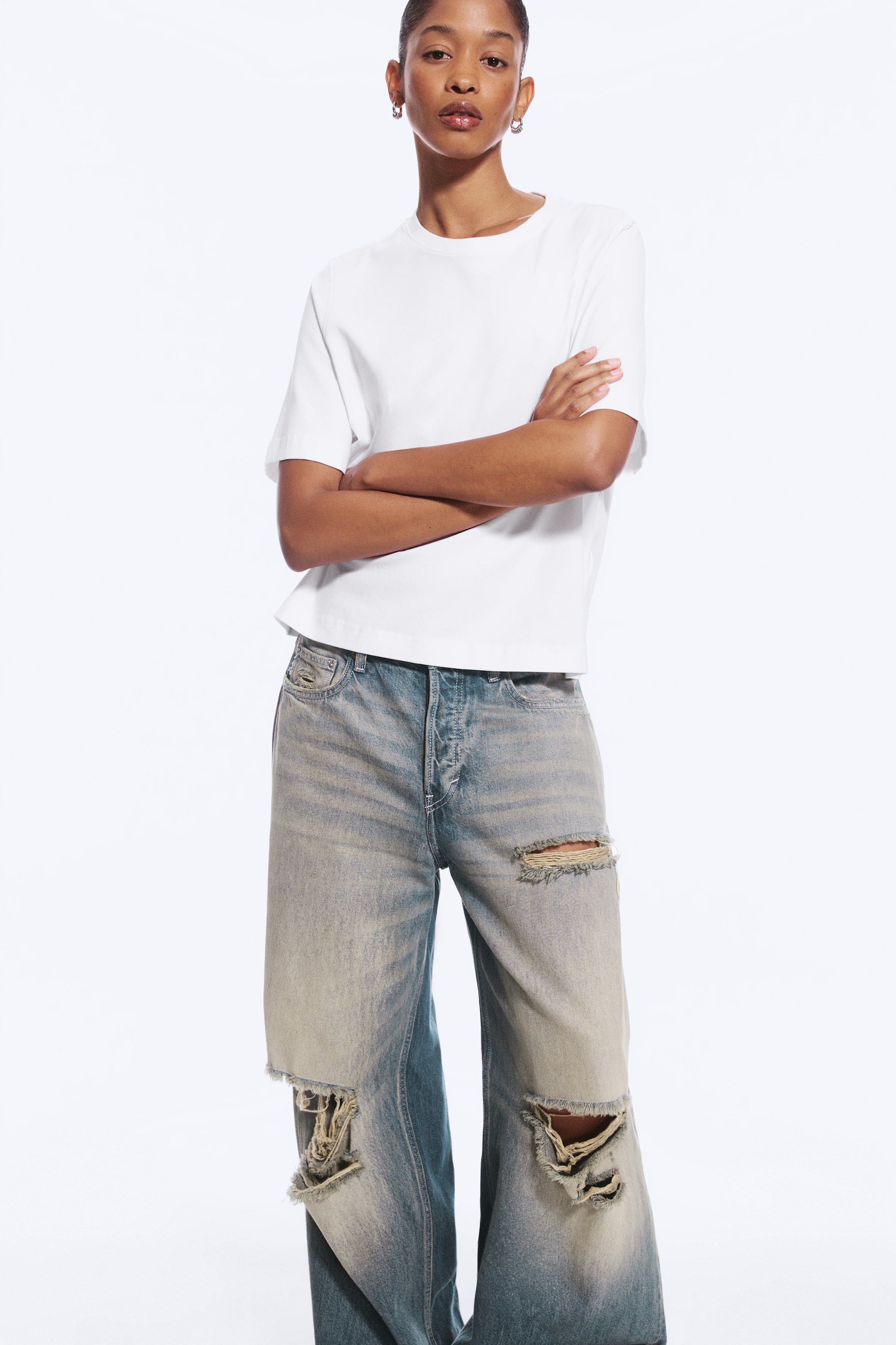 Agrandir l'image: Un t-shirt blanc oversize à col rond et manches jusqu’aux coudes est porté par le mannequin, qui se tient debout, les bras croisés. Le t-shirt est assorti à un jean bleu large délavé et usé.