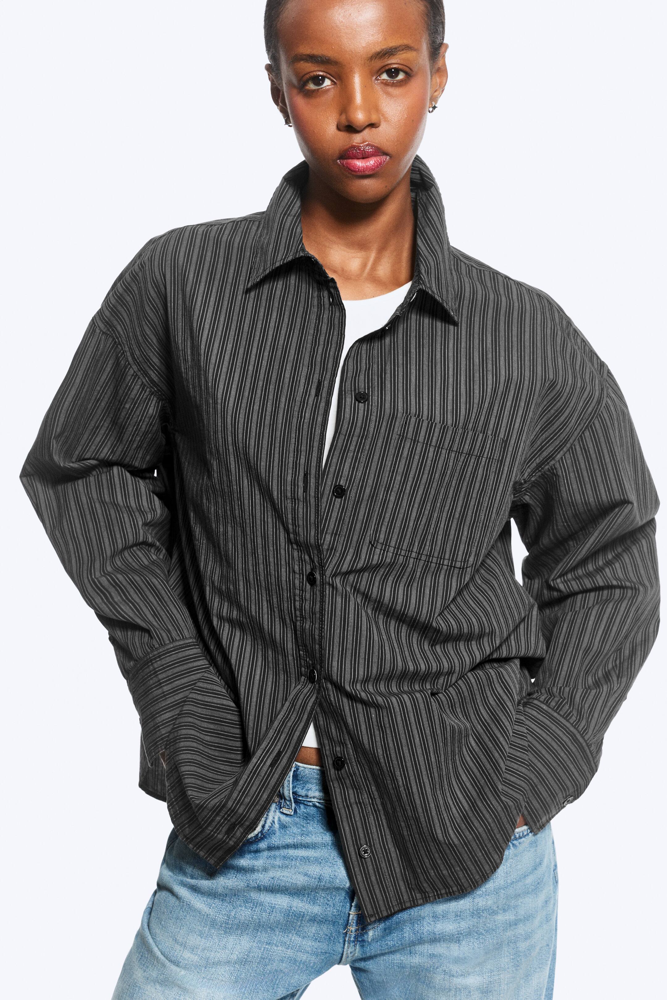 Se større billede: En mørkegrå, lodretstribet oversized skjorte bæres af modellen over en hvid T-shirt og blå jeans. Skjorten har en nedfoldet krave, knaplukning foran, en enkelt brystlomme og lange ærmer med brede manchetter. Modellen står med hænderne i jeanslommerne og fremviser skjortens afslappede, uindstoppede pasform.