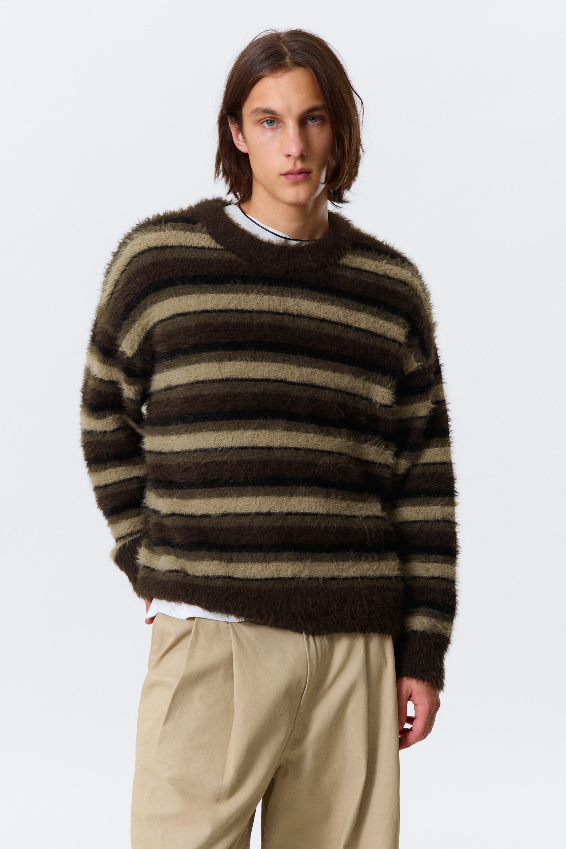 Visualizza immagine più grande: Un maglione lavorato a maglia viene presentato dalla parte anteriore, caratterizzato da una texture morbida e spazzolata e da una vestibilità rilassata con spalle scese. Presenta strisce orizzontali in tonalità di marrone scuro, marrone medio e verde oliva, con bordi a coste sul girocollo, sui polsini e sull'orlo. Una t-shirt bianca è visibile sotto il maglione.