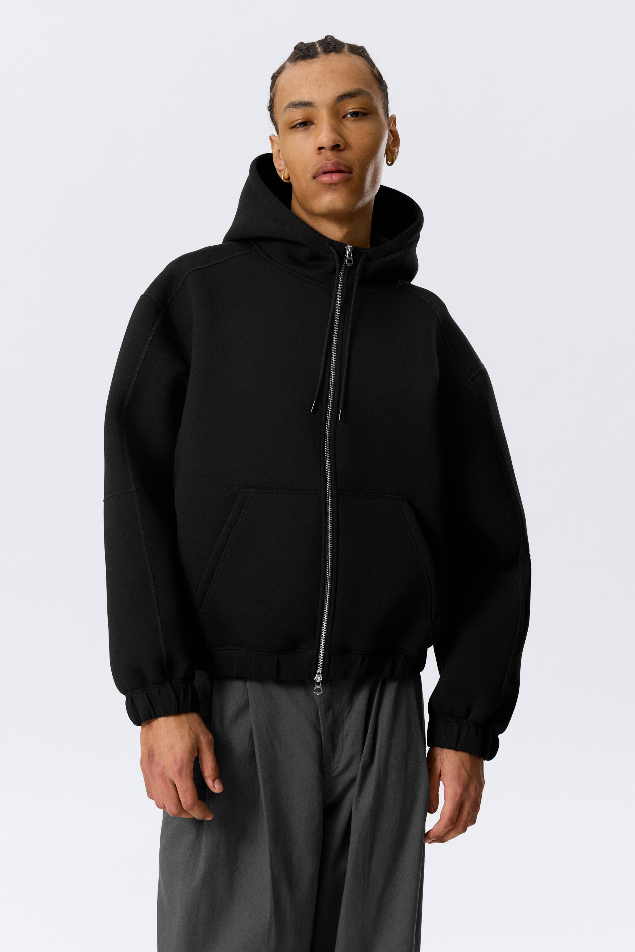 View larger image: A black full-zip hoodie, crafted from a smooth, structured fabric, is displayed from the front. It features a structured hood, a silver-toned two-way zip, and two front patch pockets. The hoodie showcases dropped shoulders, gathered cuffs, and a gathered hem, creating an oversized fit.