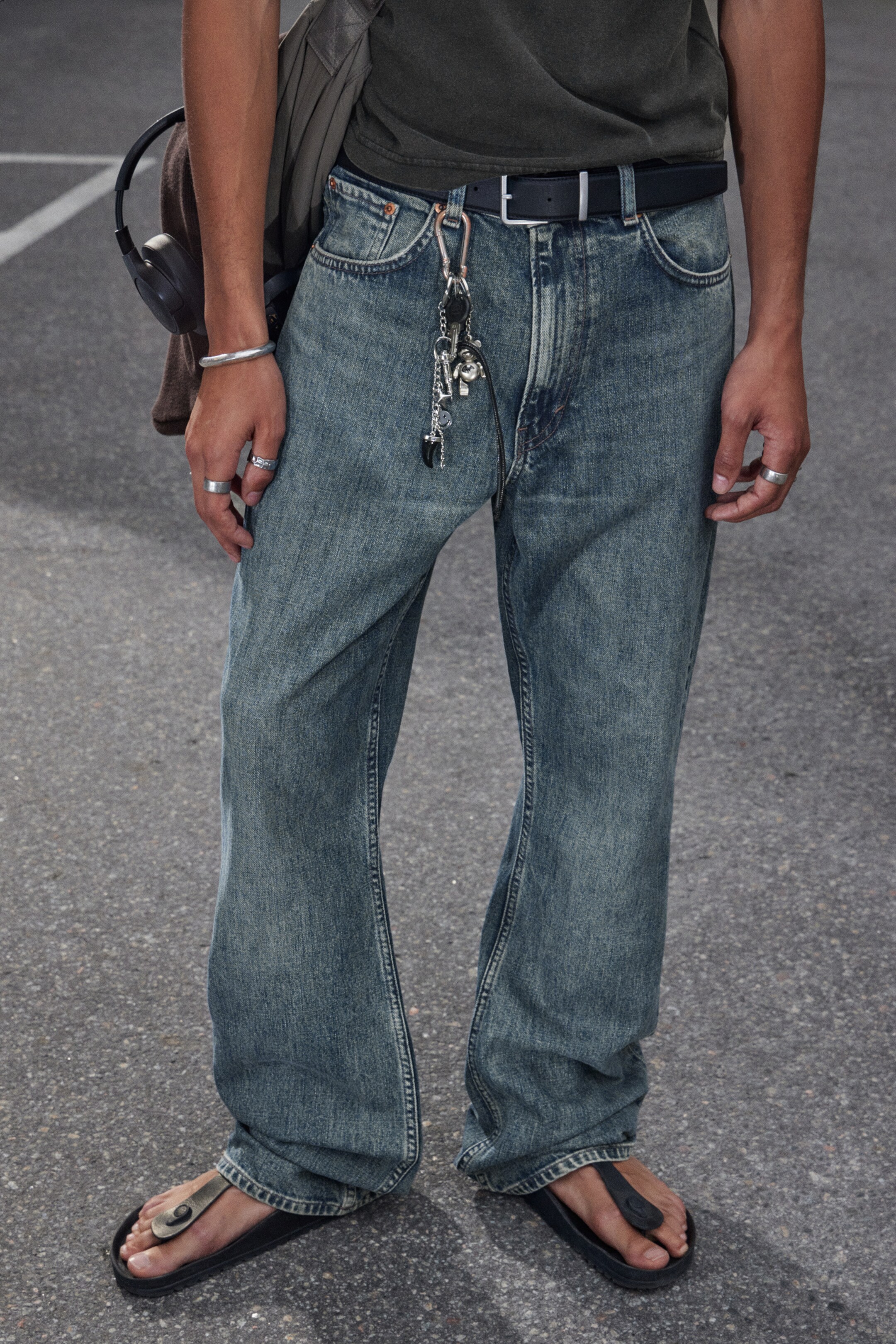 Visualizza immagine più grande: Un paio di jeans blu sbiaditi vengono mostrati da un'angolazione frontale, caratterizzati da una vestibilità rilassata con leggeri segni di usura e tasche frontali visibili. Una cintura nera stringe la vita a vita media, da cui pende un grande portachiavi con più ciondoli, e gli orli rilassati poggiano liberamente su sandali infradito neri.