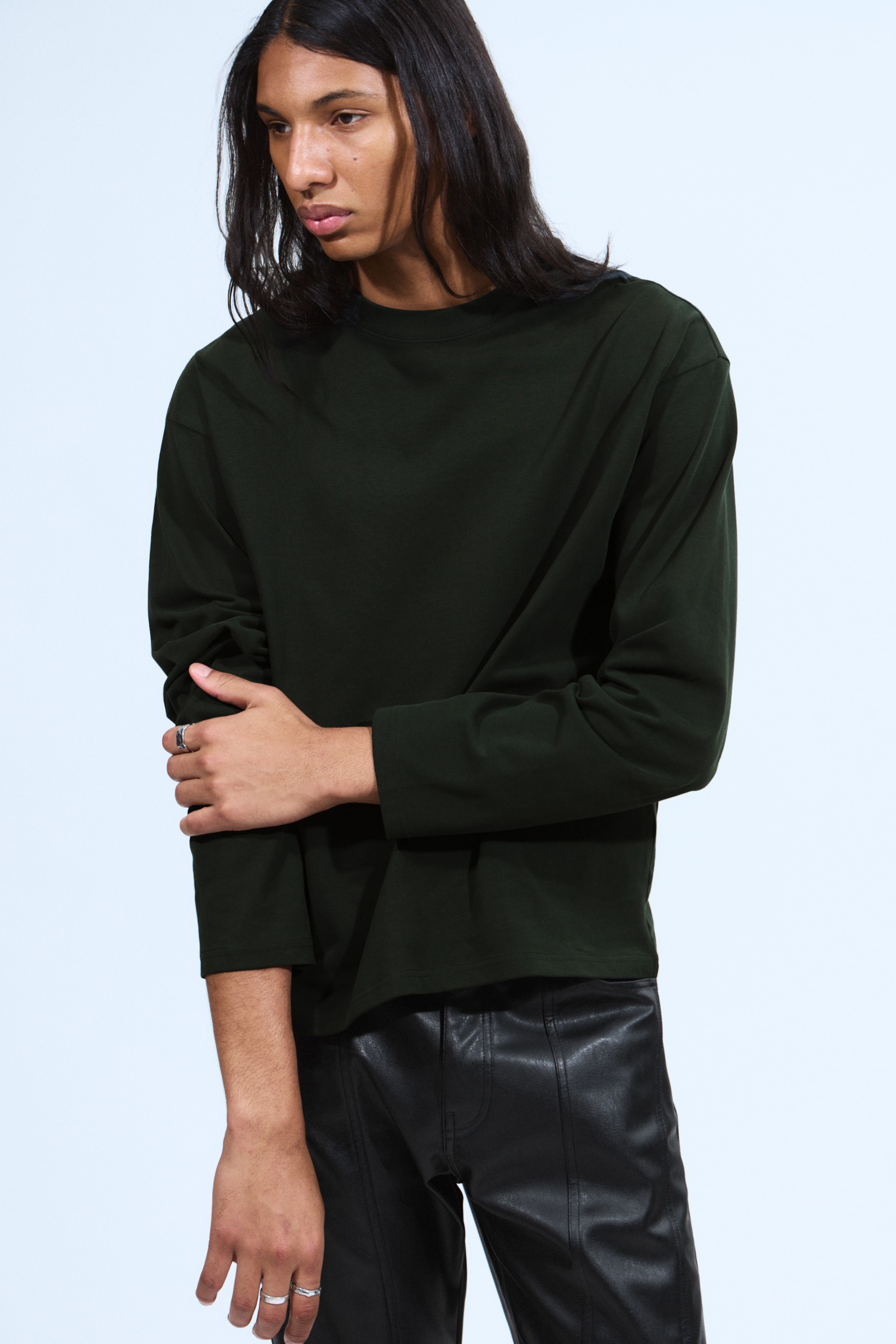 View larger image: A dark green long-sleeved shirt is being worn, featuring a crew neck and a relaxed, slightly oversized fit with straight sleeves. Black leather-look pants are also being worn, showcasing prominent vertical seams down the front of each leg.