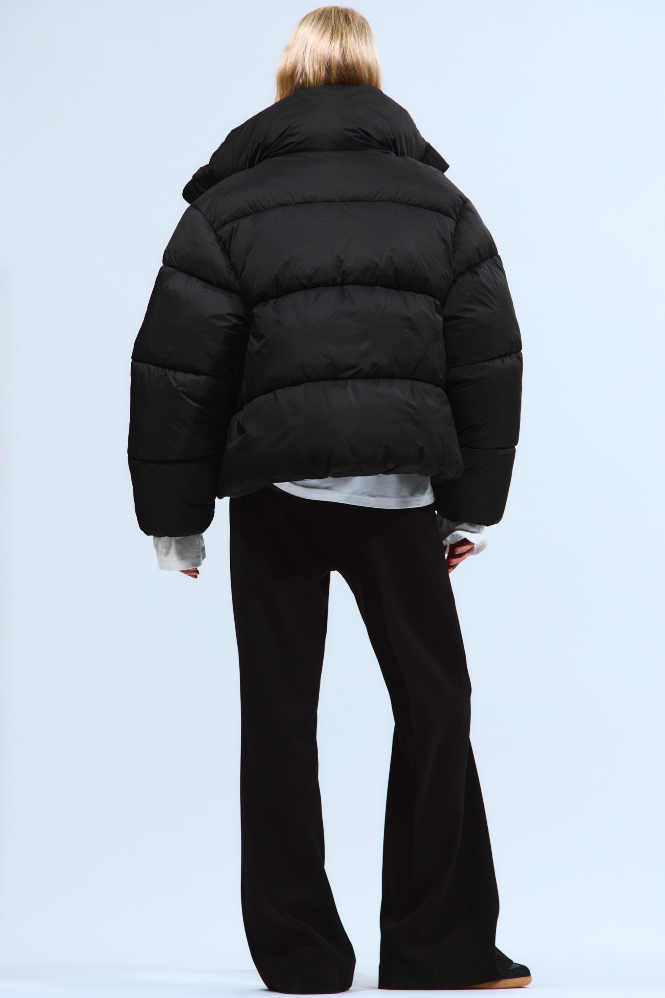 Ingrandisci l'immagine: Una giacca a vento nera viene presentata dal retro, mostrando una silhouette voluminosa e corta con cuciture orizzontali e un colletto alto. I polsini della camicia bianca sono visibilmente estesi da sotto le sue maniche lunghe.