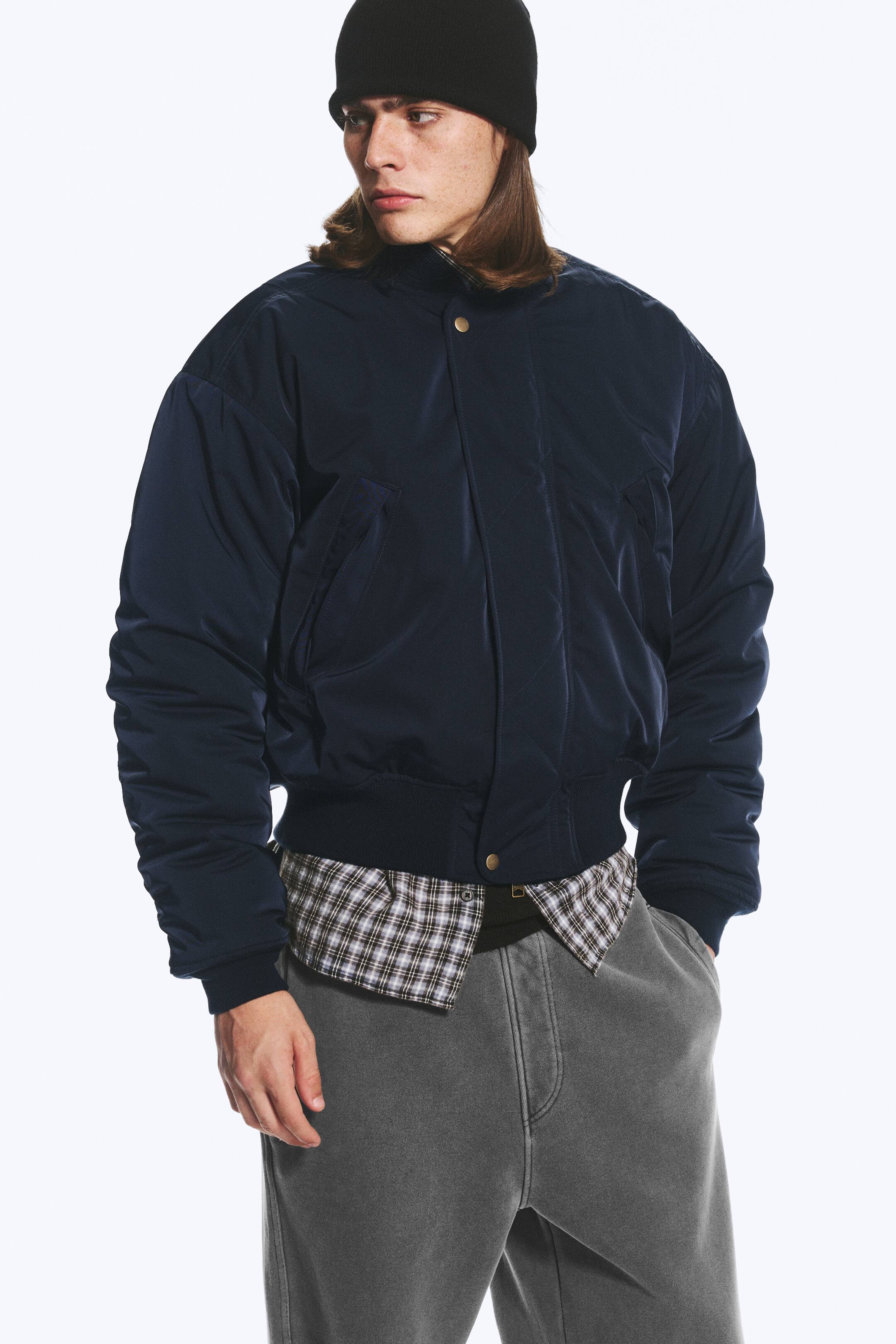 View larger image: A dark navy relaxed bomber jacket, featuring a ribbed collar, cuffs and hem, and a front placket with a visible snap fastening, is worn by the model. The jacket has a slightly lustrous finish and is layered over a plaid shirt.