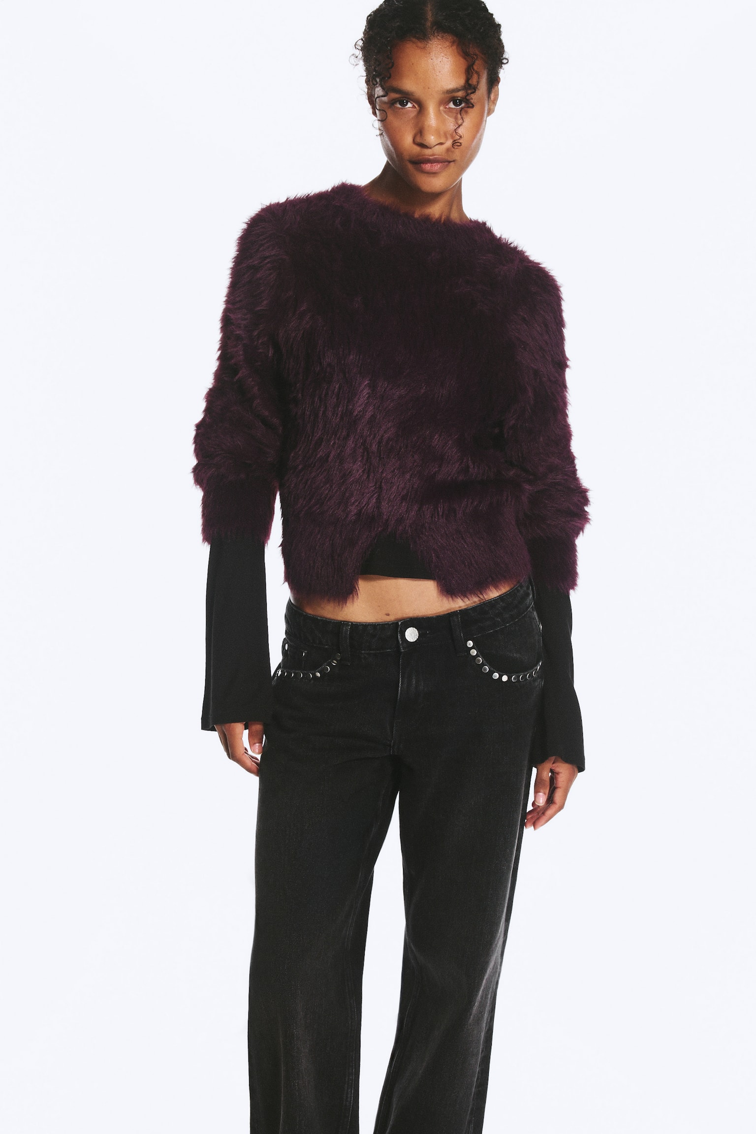 Een fluffy, donkerpaarse gebreide trui met een cropped silhouet en een split middenvoor aan de zoom wordt gedragen door het model. De tactile trui met ronde hals is gelaagd over een zwarte top met lange mouwen en gecombineerd met zwarte jeans. - 1
