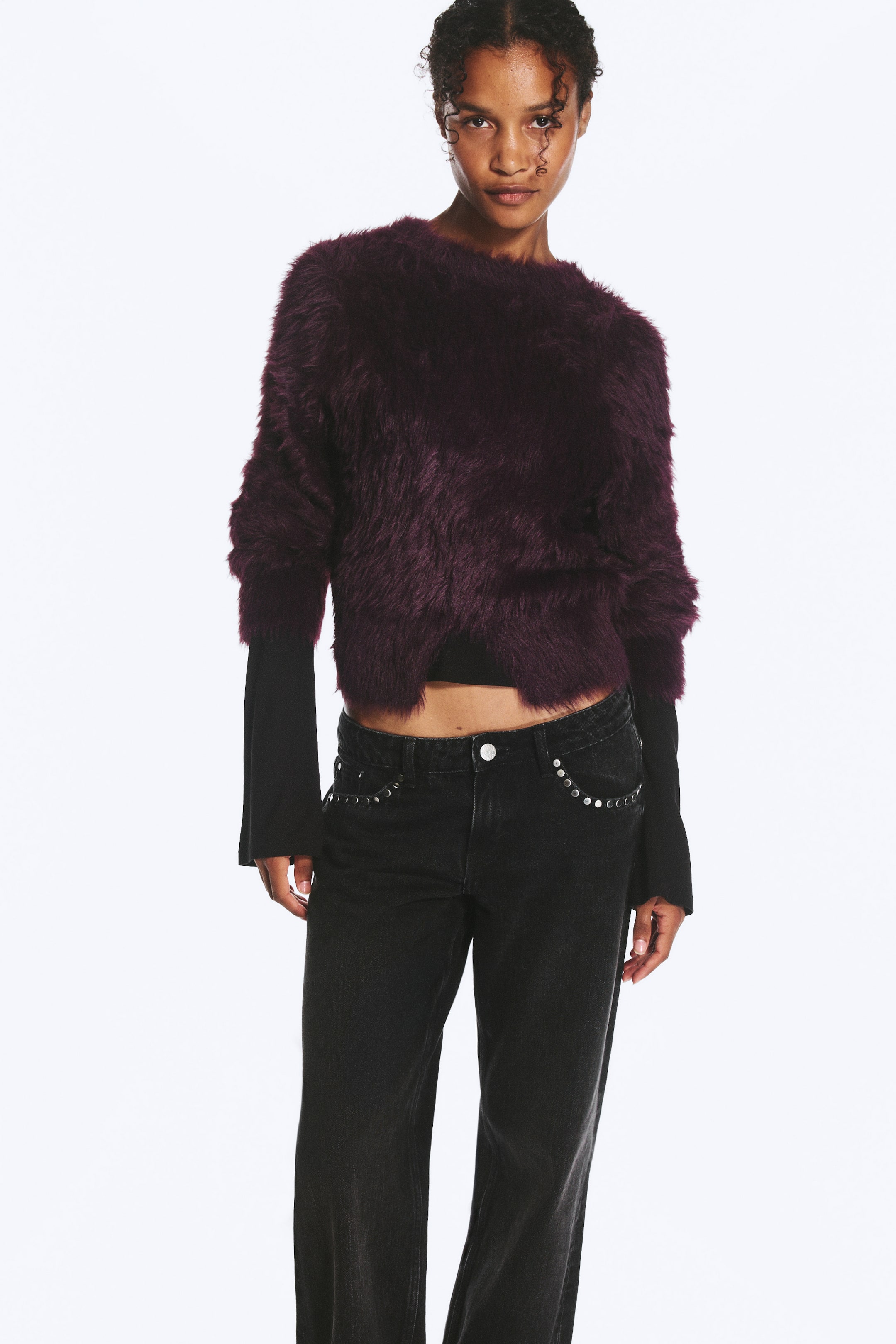 Größeres Bild ansehen: Ein flauschiger, dunkelvioletter Strickpullover mit cropped Silhouette und einem mittigen Schlitz am Saum wird von dem Model getragen. Der griffige Pullover mit Rundhalsausschnitt wird über einem schwarzen Langarm-Top getragen und mit schwarzen Jeans kombiniert.