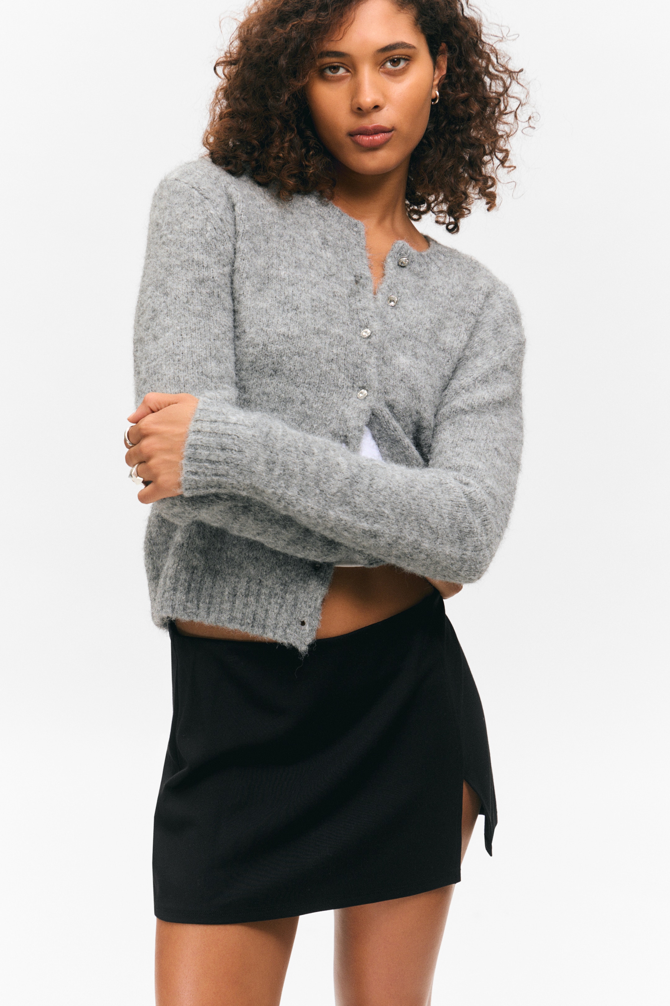 Größeres Bild ansehen: Eine graue flauschige Strickjacke wird getragen, mit einem Rundhalsausschnitt, langen Ärmeln mit gerippten Bündchen und einem gerippten, verkürzten Saum. Sie wird vorne mit klaren Schmuckknöpfen geschlossen und über einem weißen Oberteil kombiniert, während auch ein schwarzer Minirock mit einem seitlichen Schlitz getragen wird.