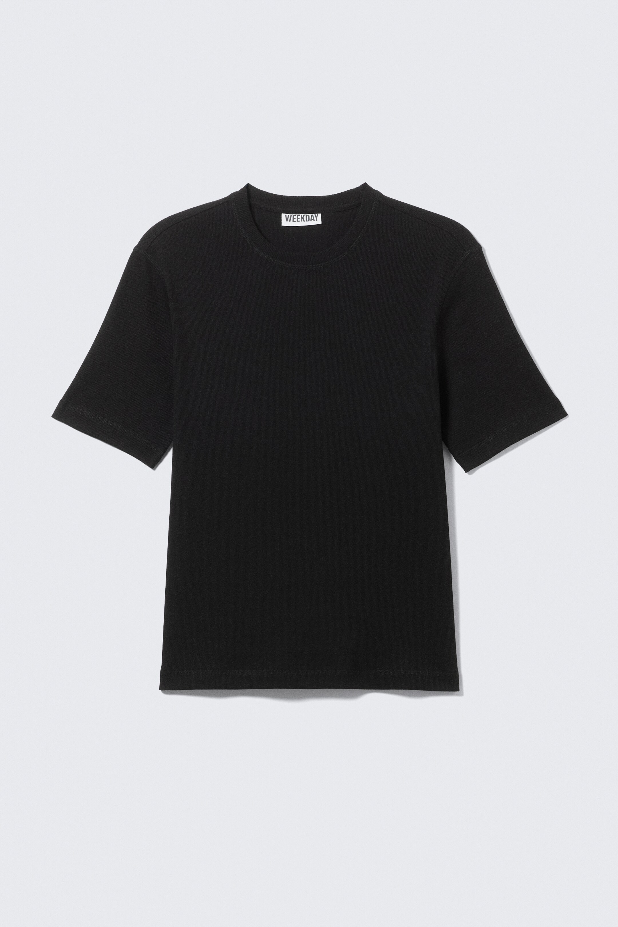 Vezi imaginea mai mare: Un tricou negru prezintă partea din față, cu o textură fină, reiată. Are guler rotund clasic și mâneci scurte, iar o etichetă de brand 'WEEKDAY' este vizibilă în partea din spate a gulerului.